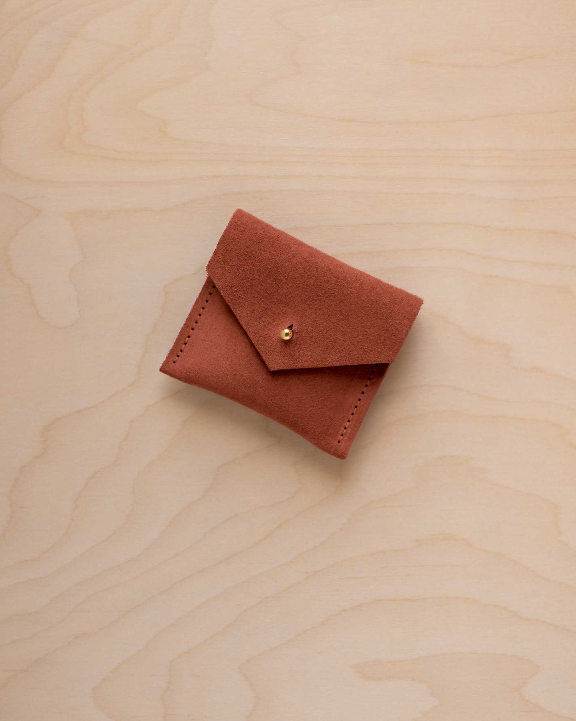 A small terracotta-colored suede pouch with a flap closure and a gold button, resting on a light wood grain surface.