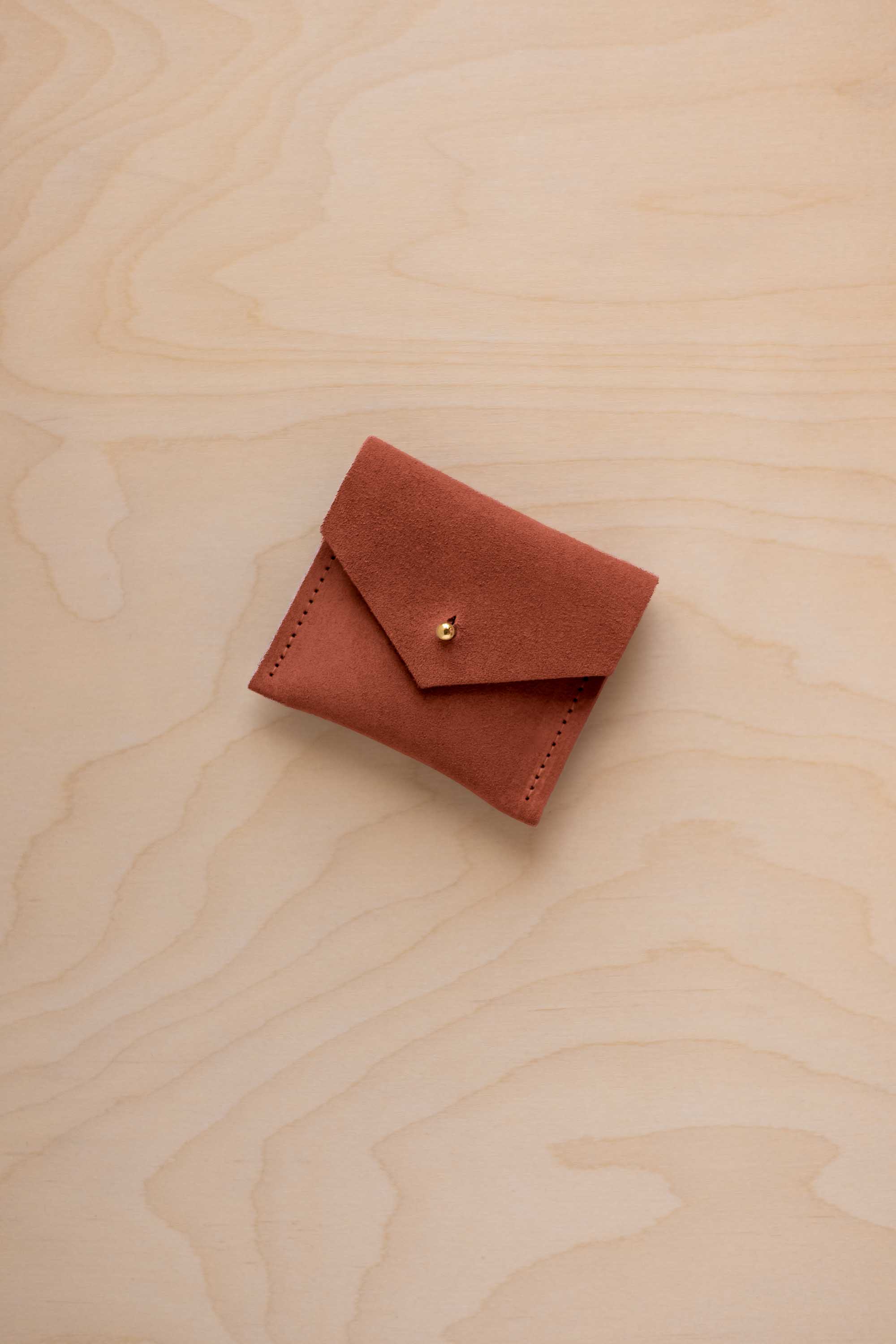 A small terracotta-colored suede pouch with a flap closure and a gold button, resting on a light wood grain surface.