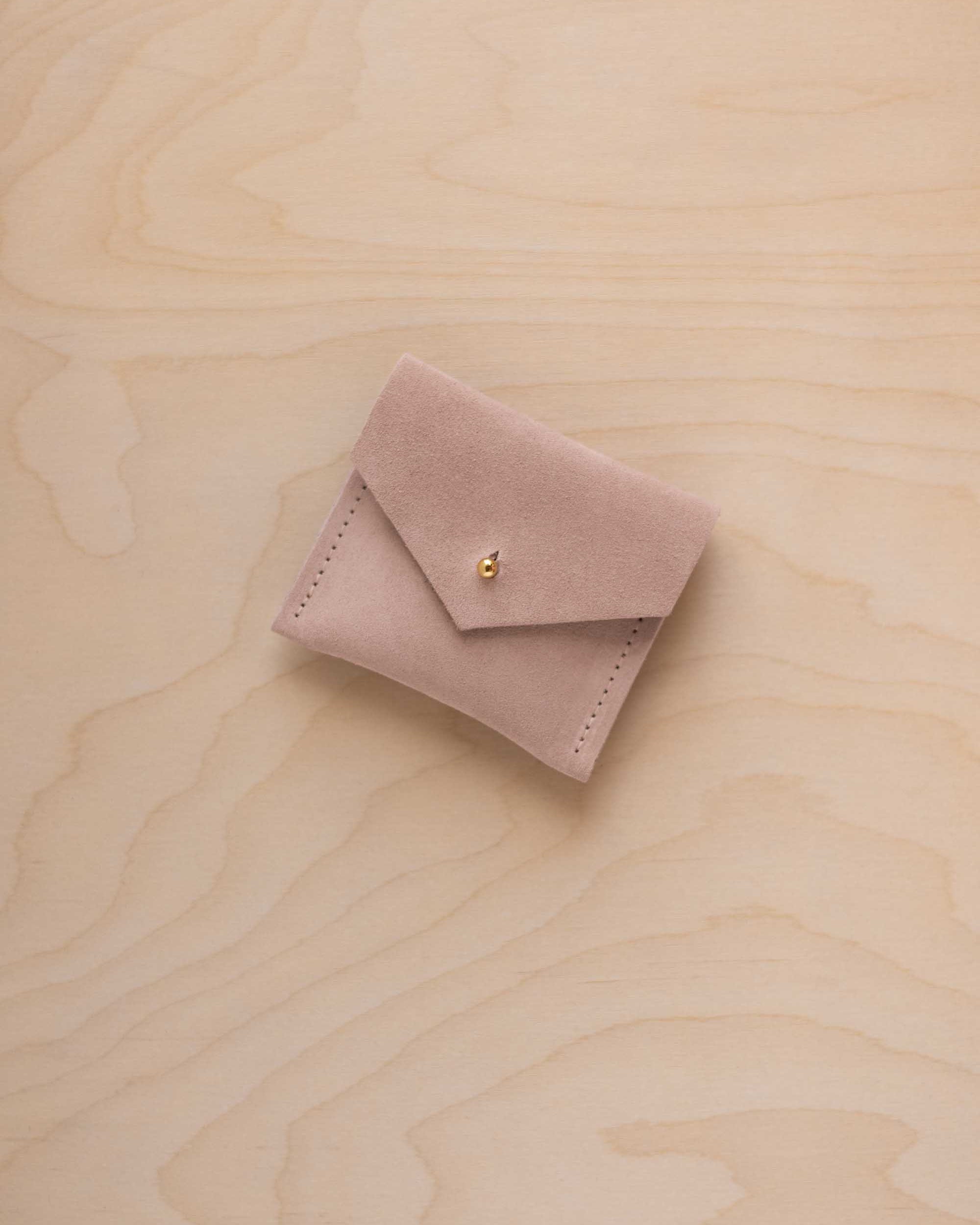A small warm beige suede jewelry pouch with a gold button, resting on a light wood grain surface.