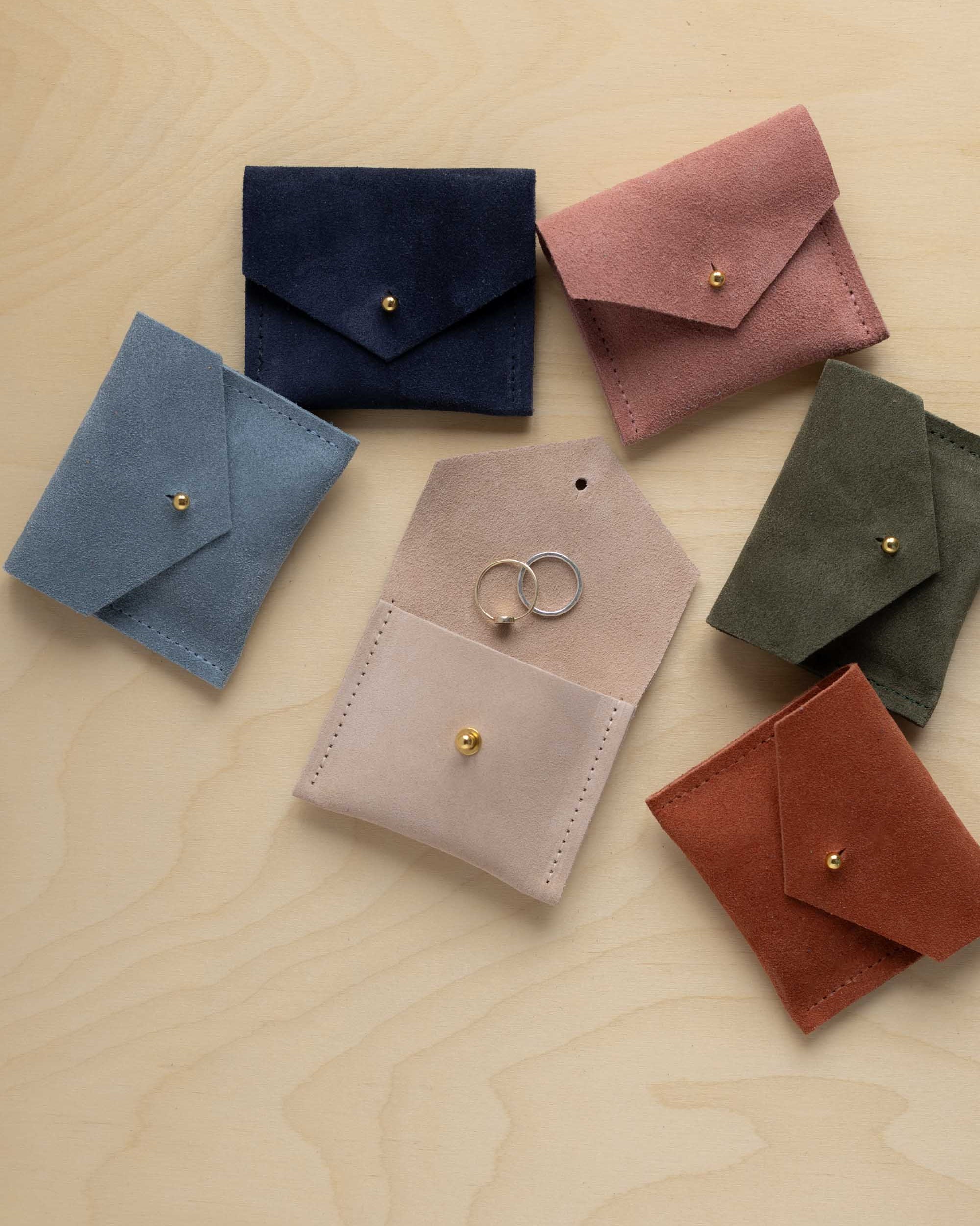 Six small suede envelope style pouches in different colours arranged on a light wood surface. The pouches are blue, navy, pink, olive green, blush, and rust, each with a small gold-tone stud closure.