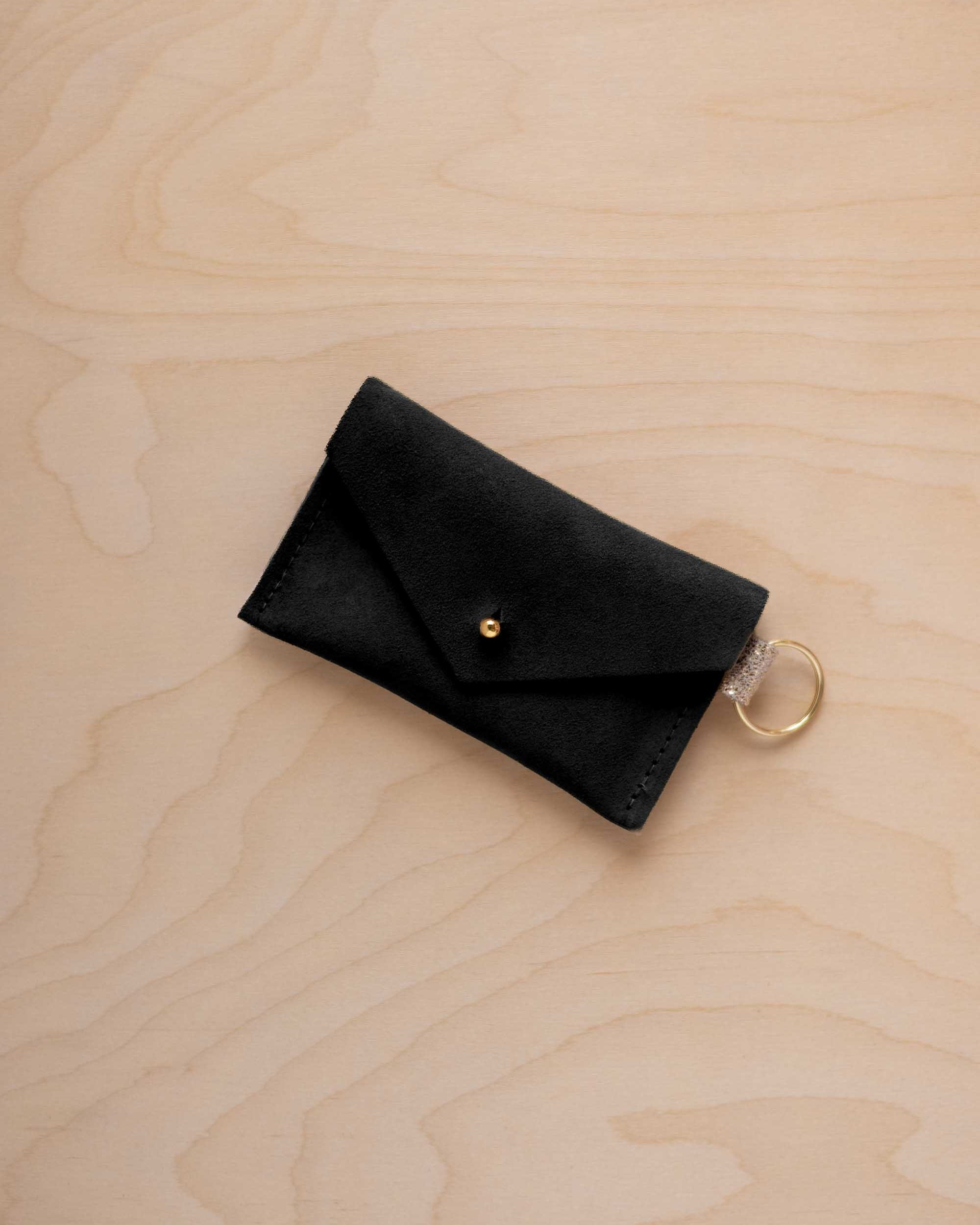 A small soft black coloured suede envelope-style keyring pouch with a gold button, resting on a light wood grain surface.