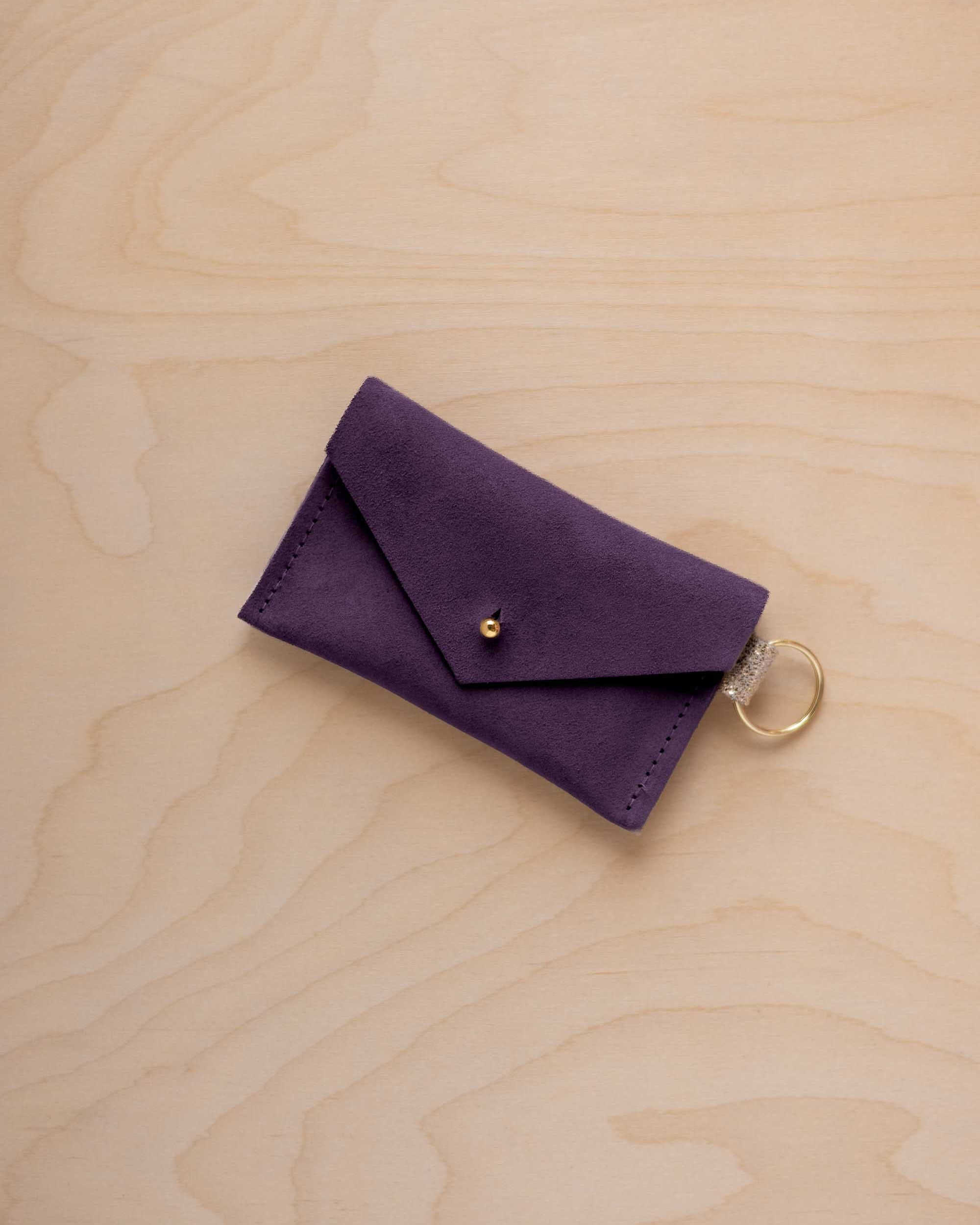 A small blackcurrant coloured suede envelope-style keyring pouch with a gold button, resting on a light wood grain surface.