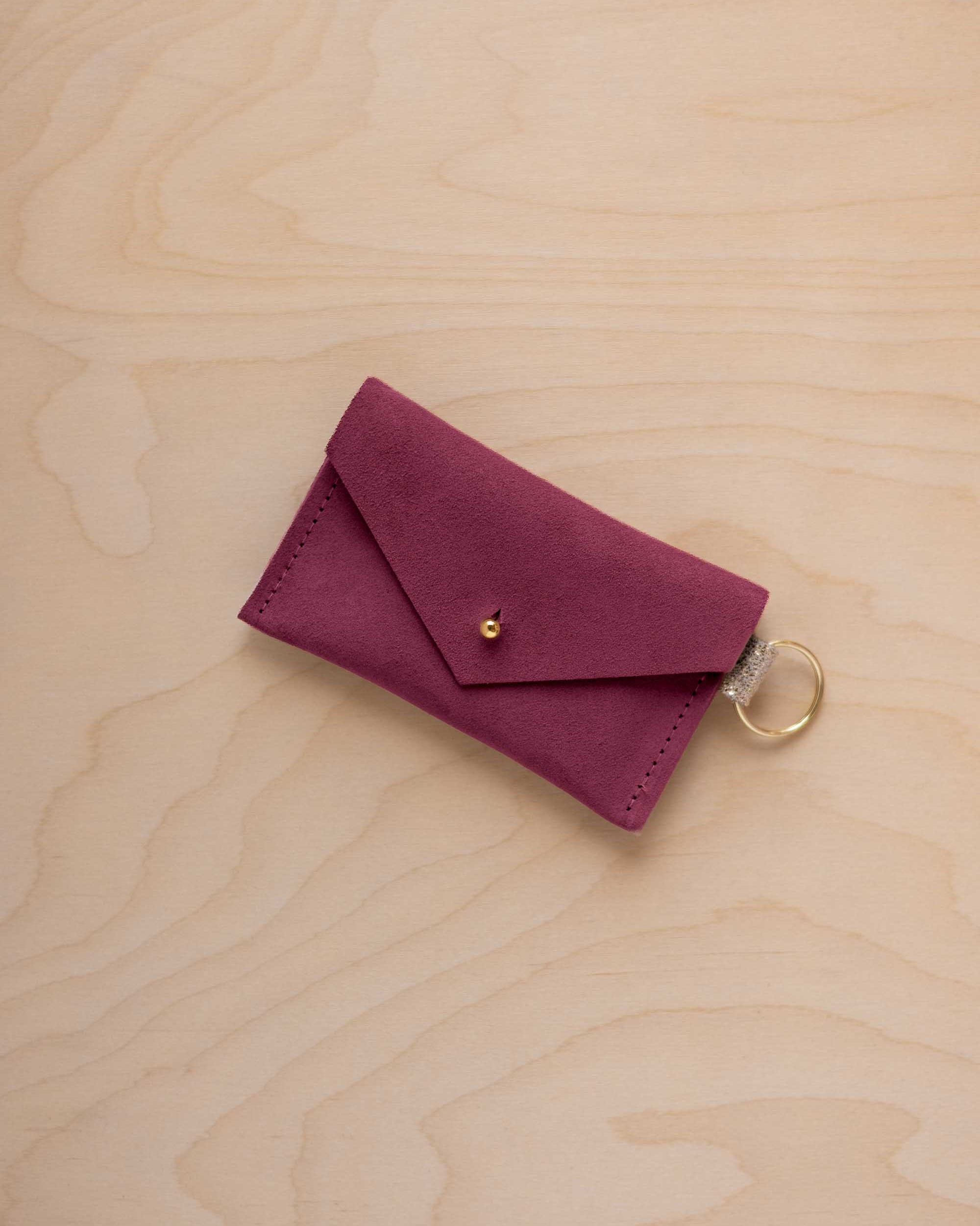 A small burgundy coloured suede envelope-style keyring pouch with a gold button, resting on a light wood grain surface.