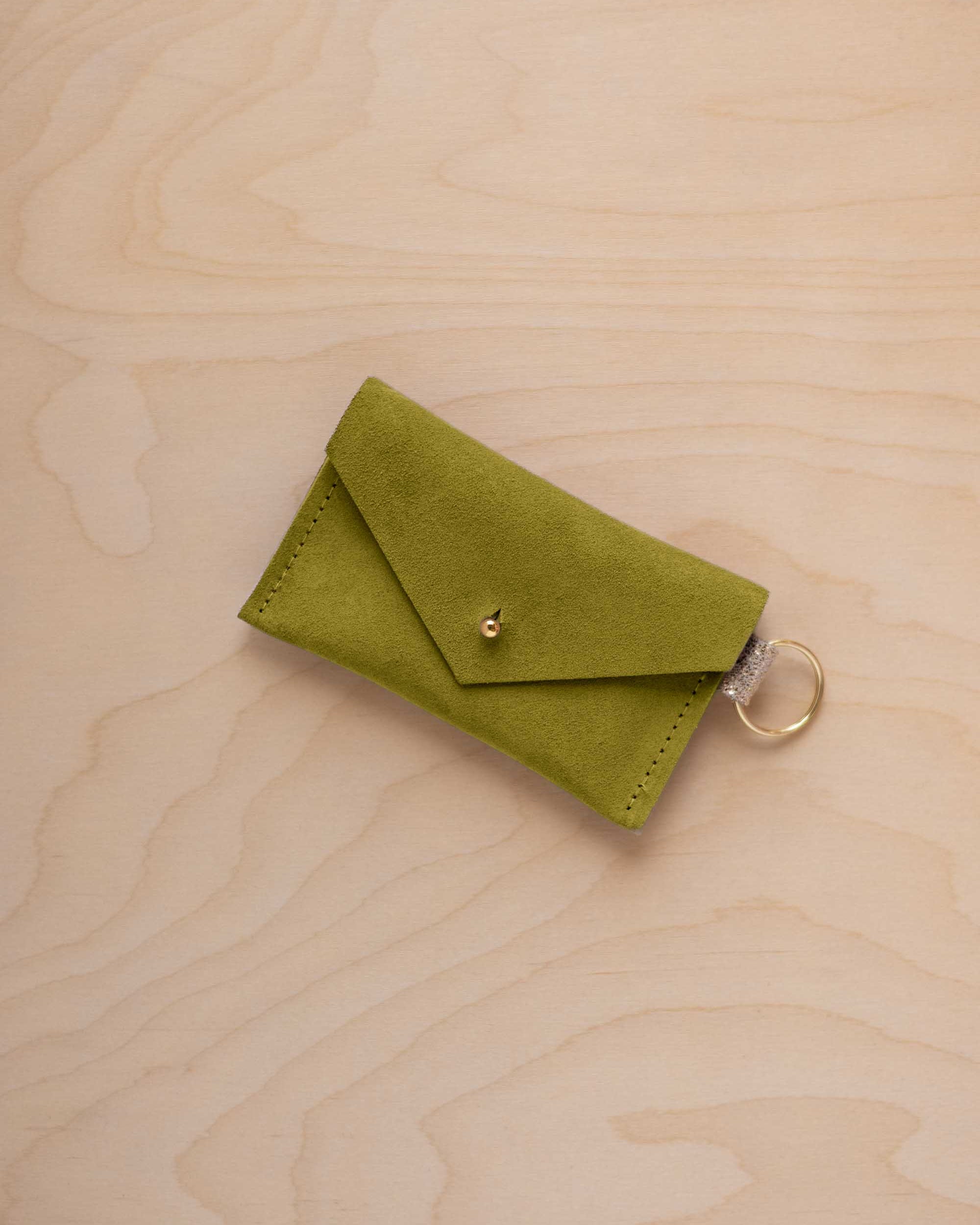 A small chartreuse green coloured suede envelope-style keyring pouch with a gold button, resting on a light wood grain surface.
