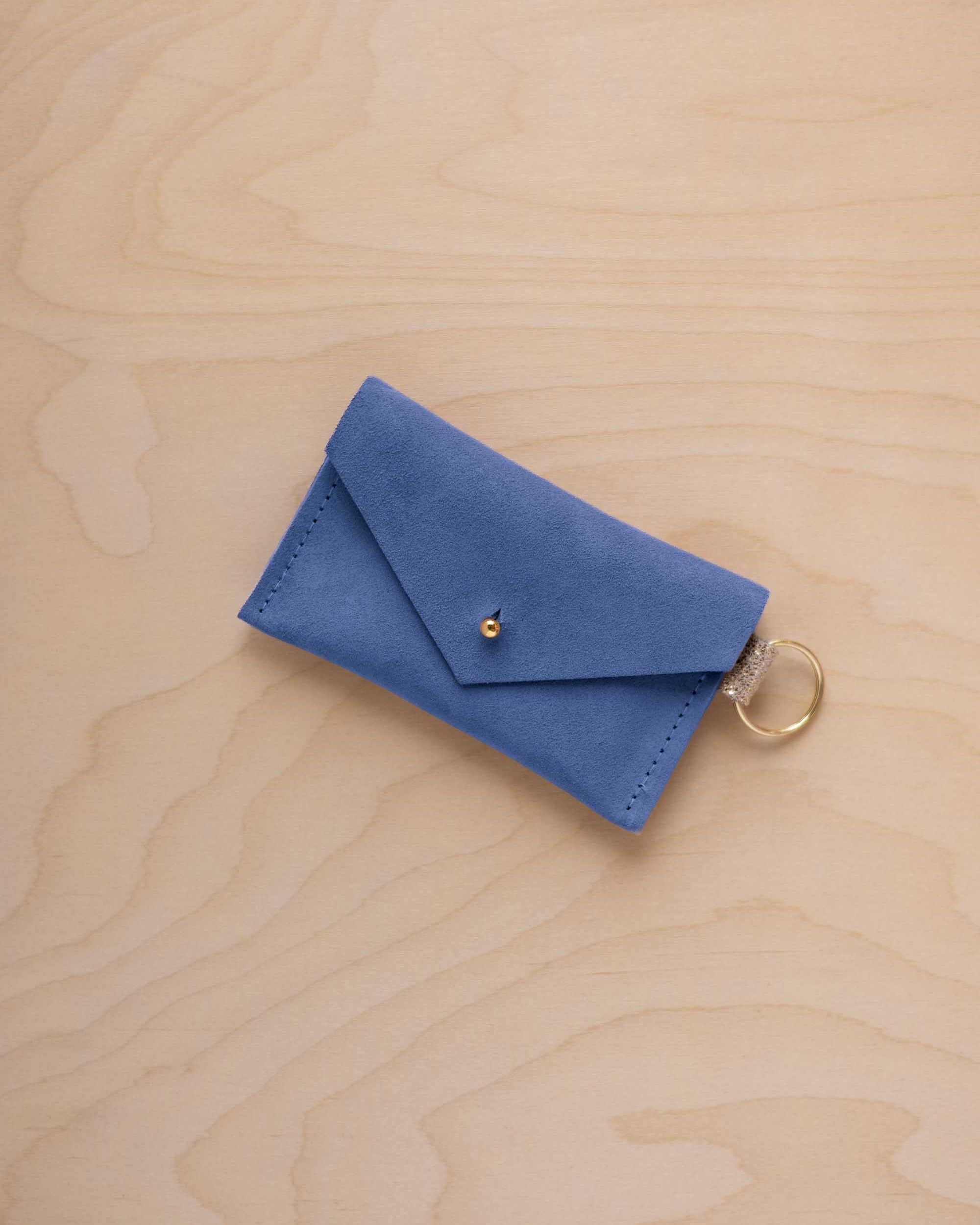A small cobalt blue coloured suede envelope-style keyring pouch with a gold button, resting on a light wood grain surface.