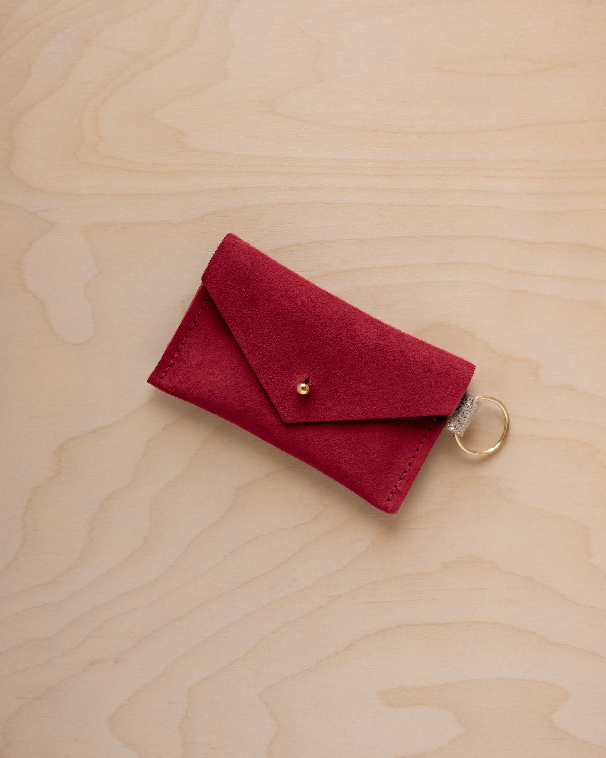 A small crimson red coloured suede envelope-style keyring pouch with a gold button, resting on a light wood grain surface.