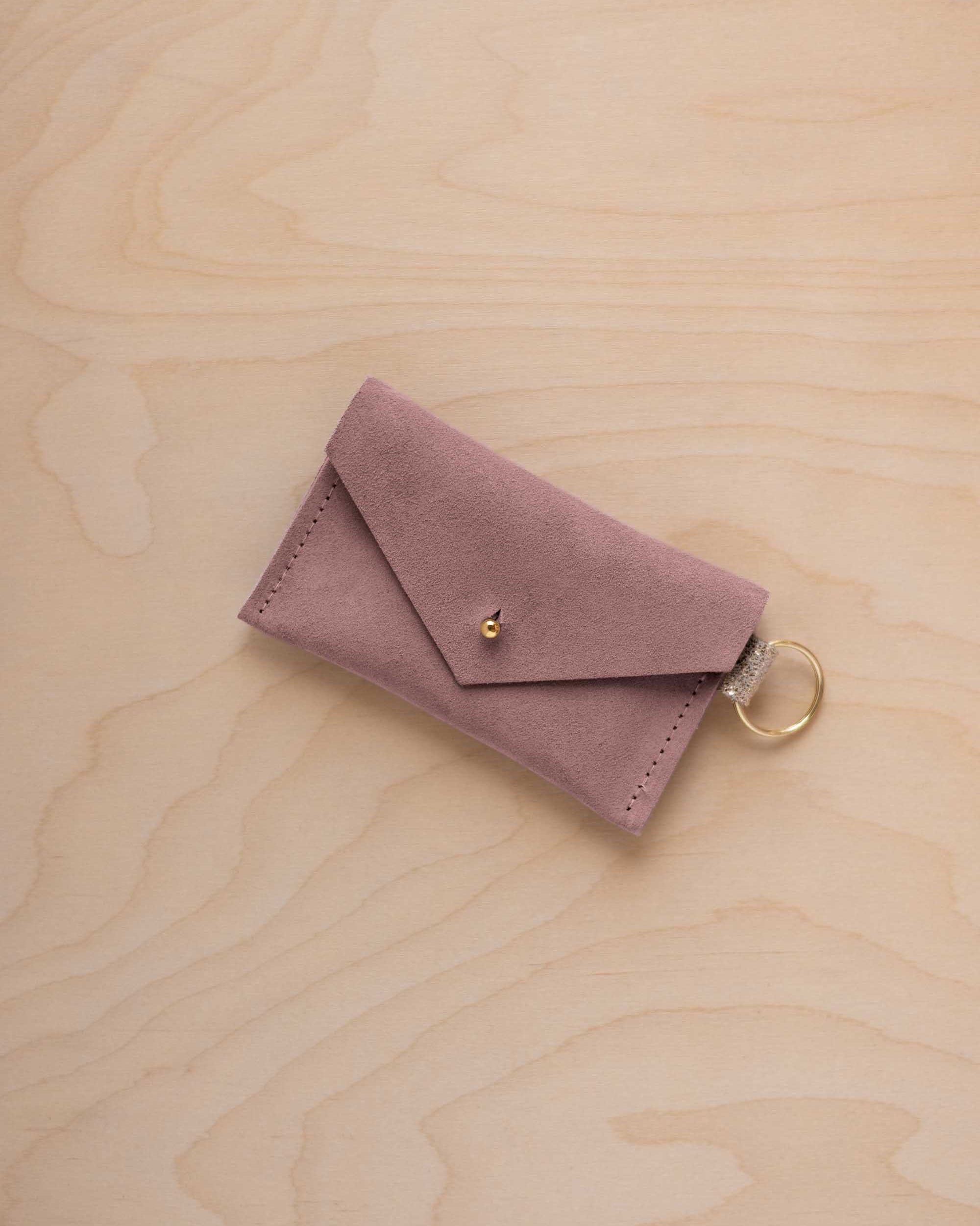 A small dusty mauve coloured suede envelope-style keyring pouch with a gold button, resting on a light wood grain surface.