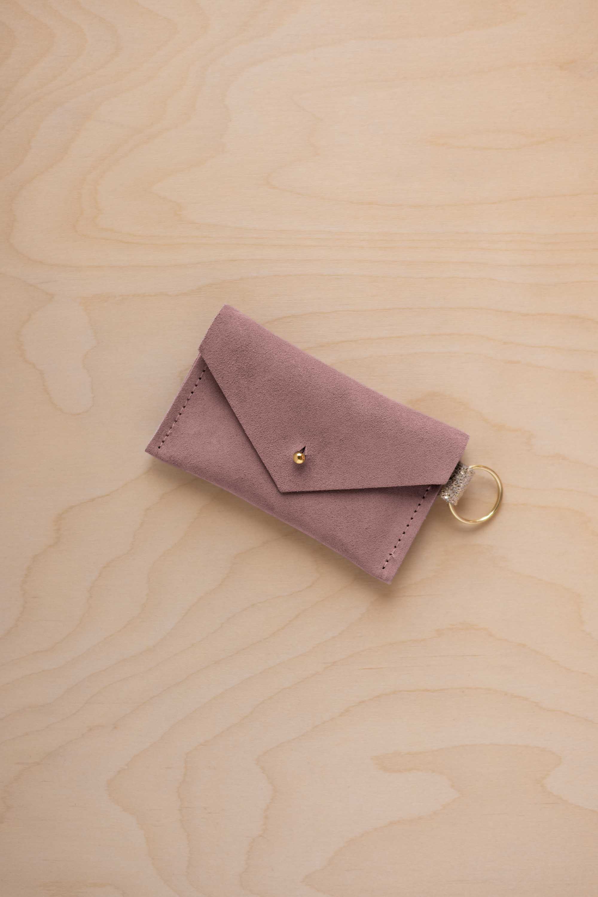 A small dusty mauve coloured suede envelope-style keyring pouch with a gold button, resting on a light wood grain surface.