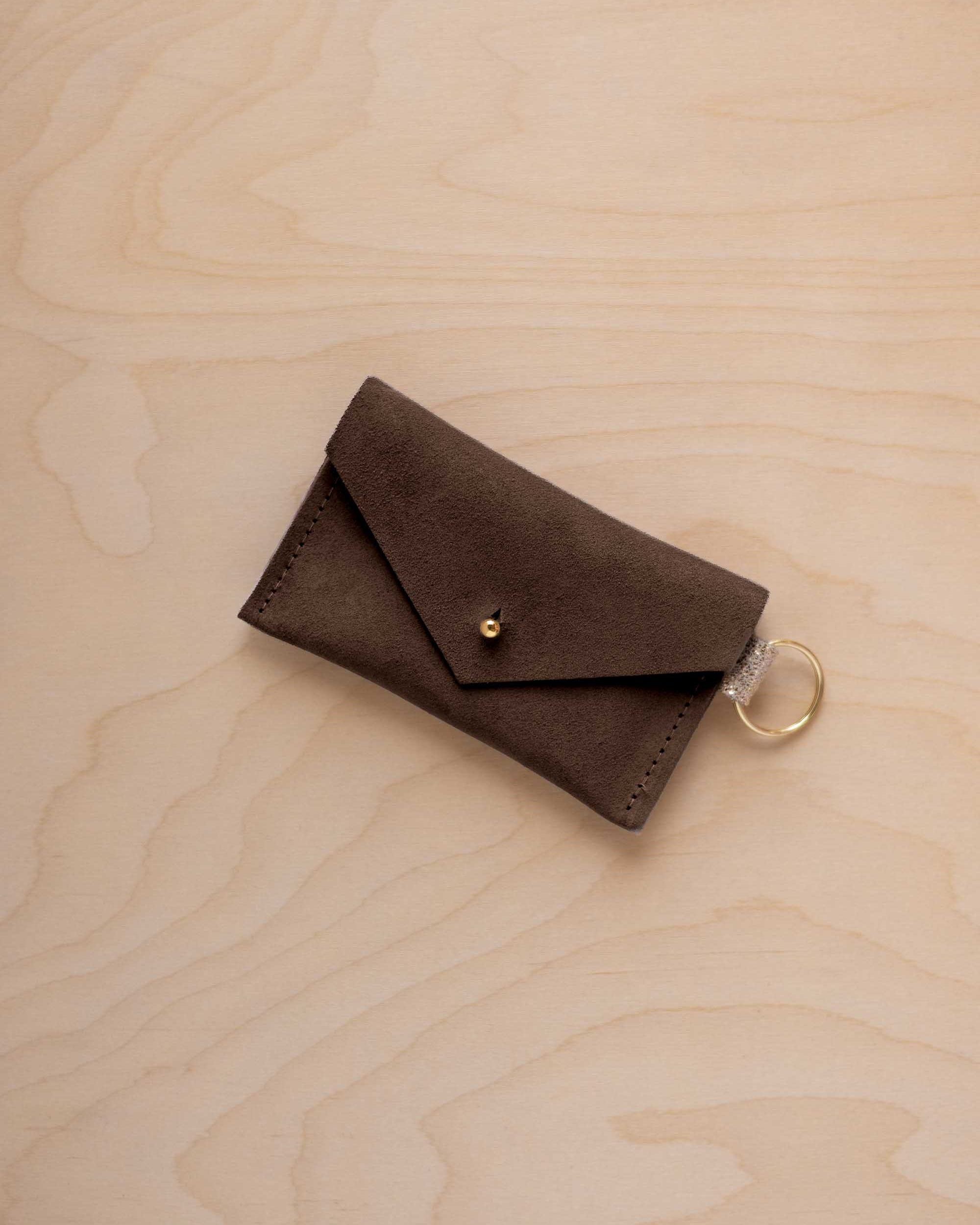 A small espresso brown coloured suede envelope-style keyring pouch with a gold button, resting on a light wood grain surface.