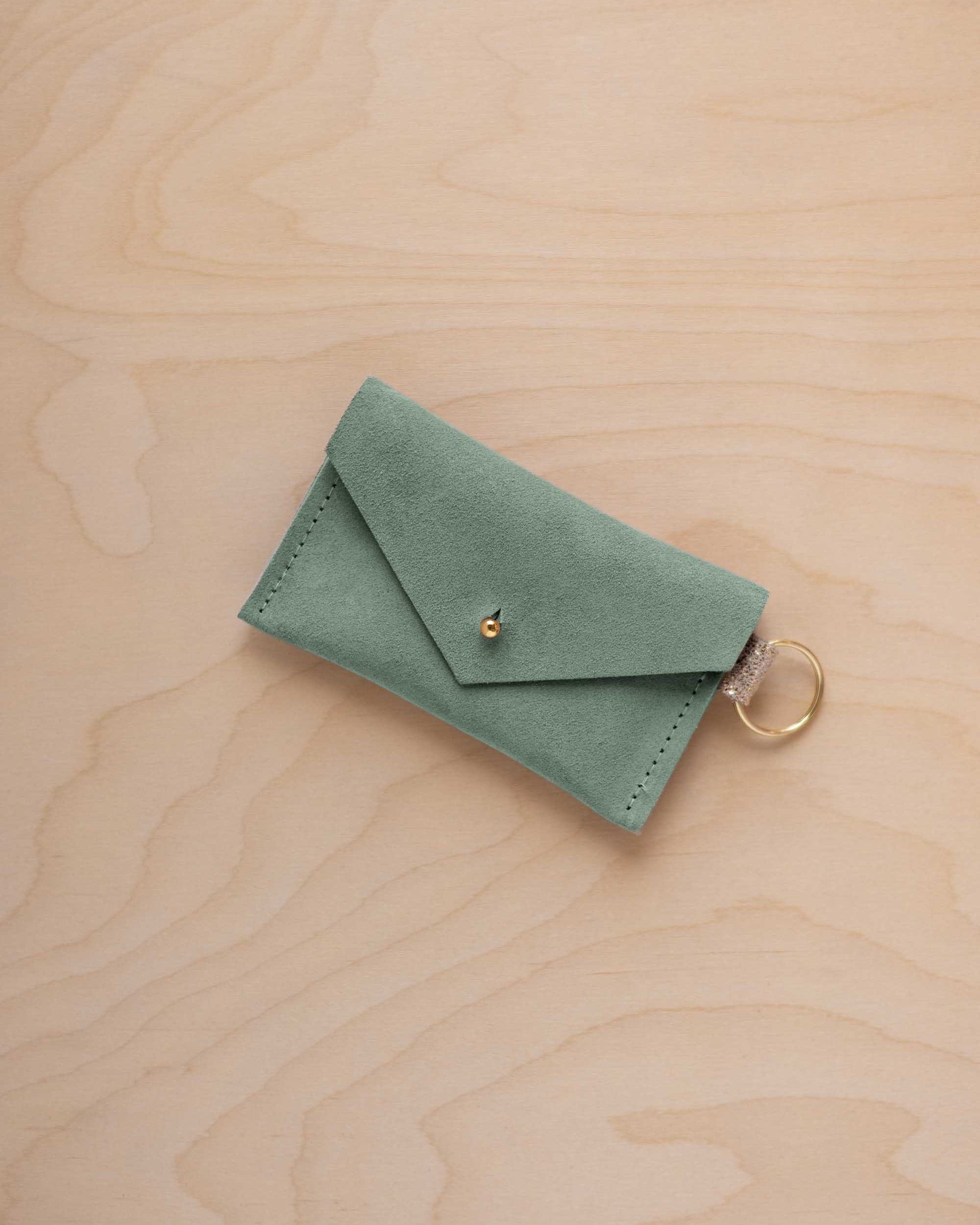 A small eucalyptus green coloured suede envelope-style keyring pouch with a gold button, resting on a light wood grain surface.