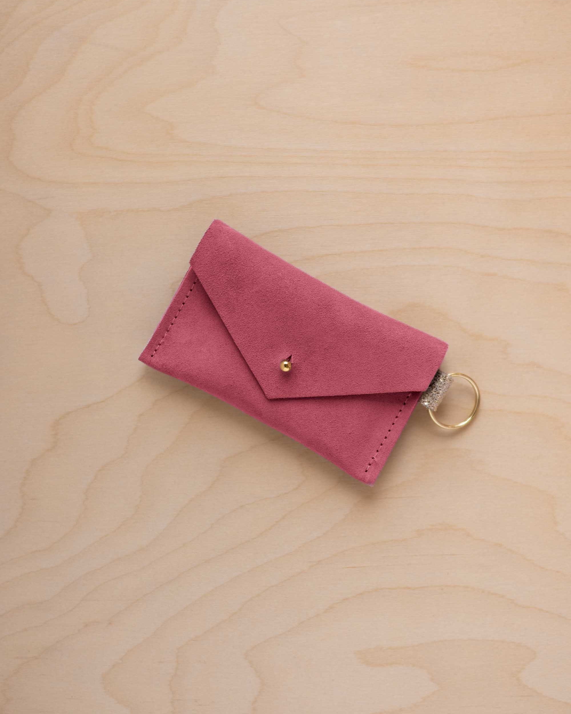 A small faded rose coloured suede envelope-style keyring pouch with a gold button, resting on a light wood grain surface.