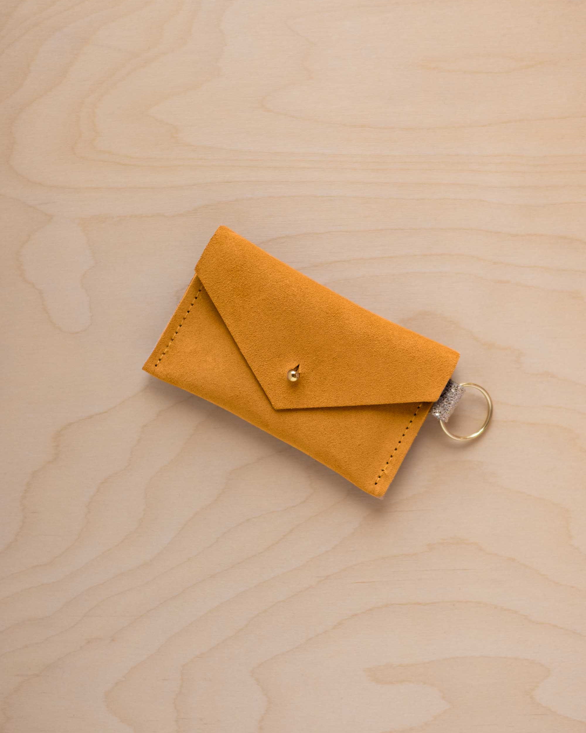 A small golden ochre coloured suede envelope-style keyring pouch with a gold button, resting on a light wood grain surface.