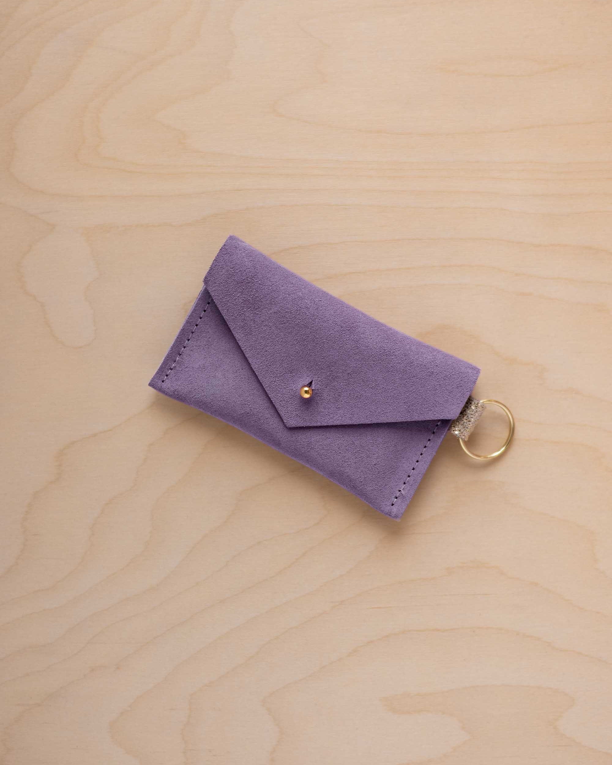 A small lavender coloured suede envelope-style keyring pouch with a gold button, resting on a light wood grain surface.