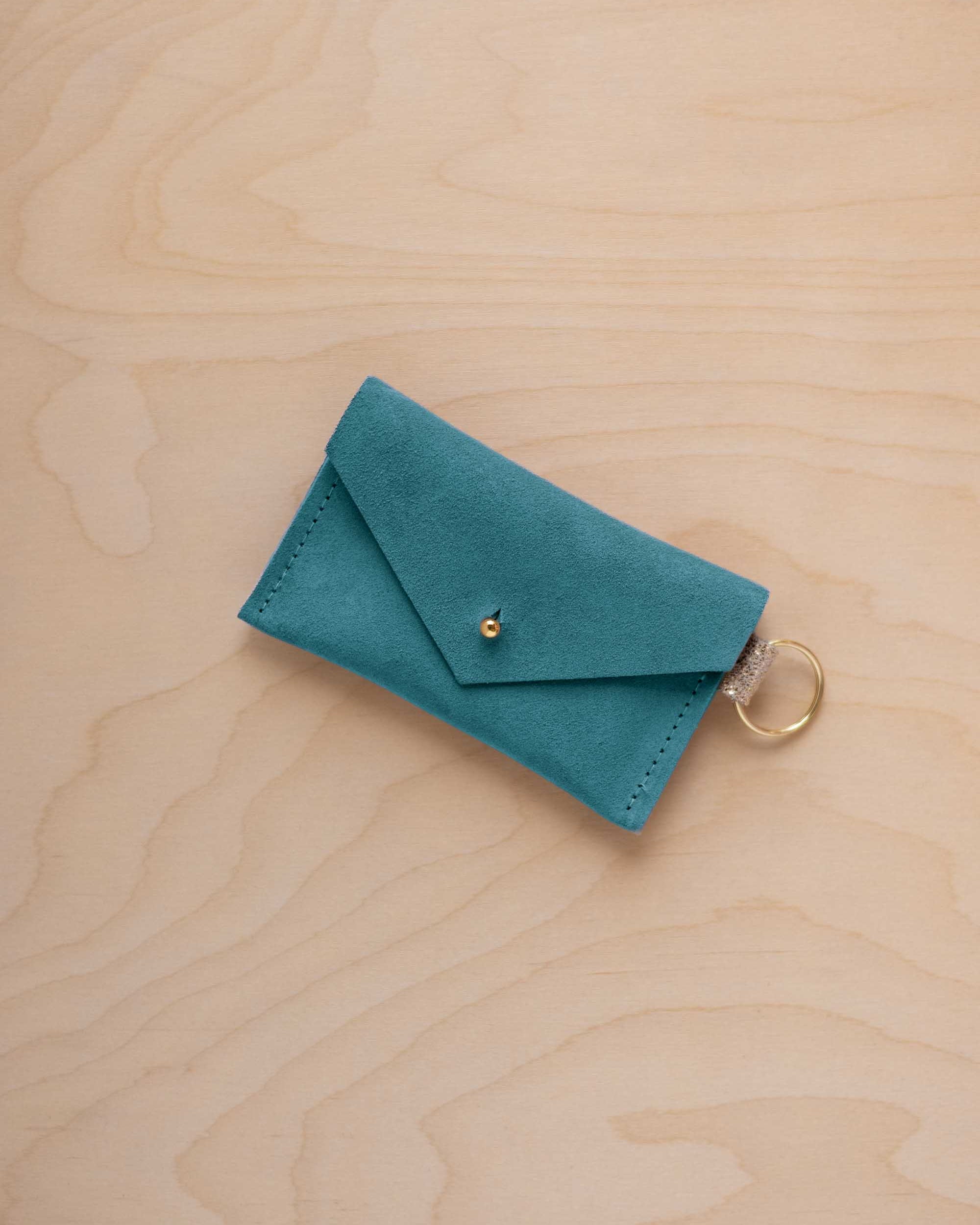 A small marine teal coloured suede envelope-style keyring pouch with a gold button, resting on a light wood grain surface.