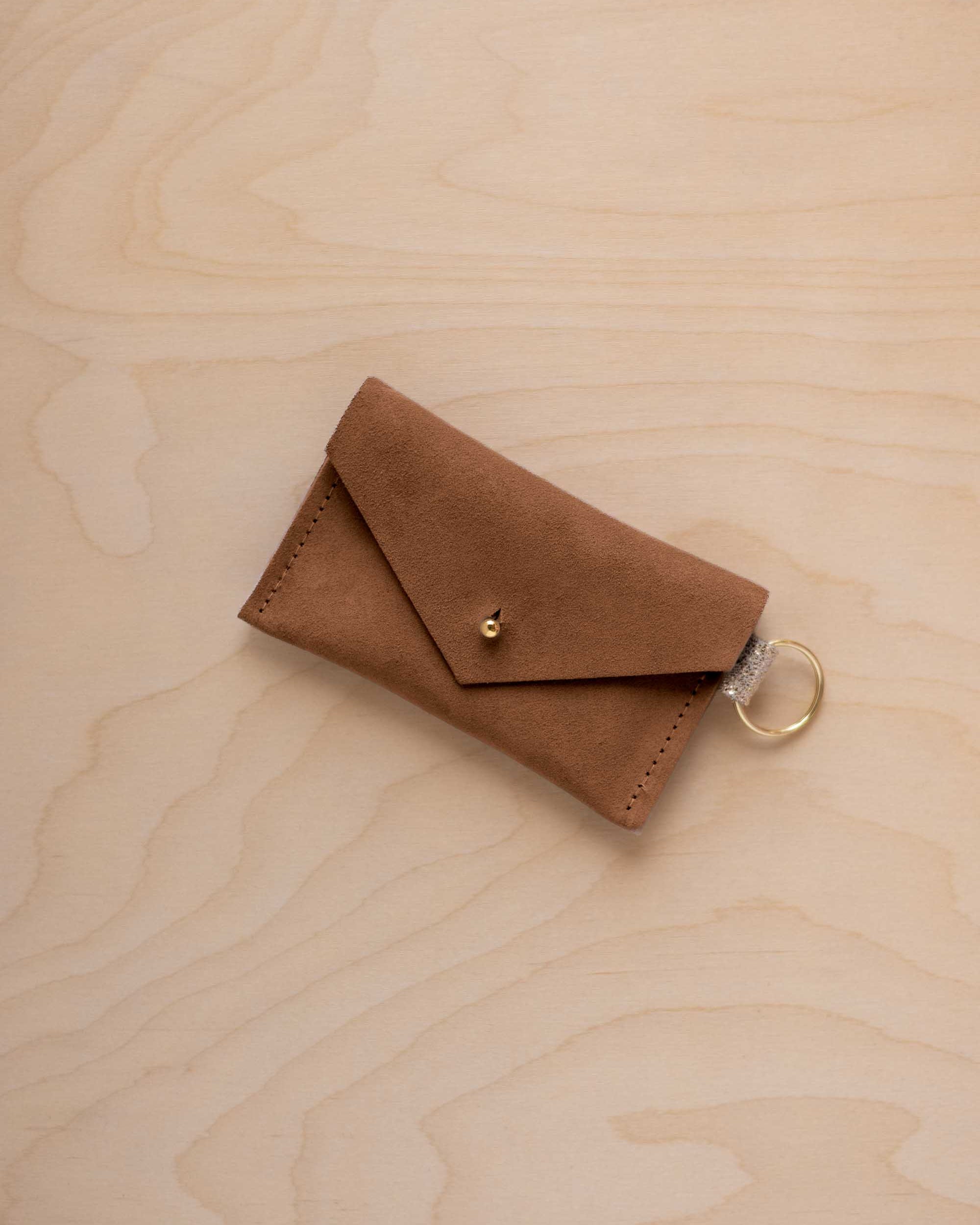A small mocha brown coloured suede envelope-style keyring pouch with a gold button, resting on a light wood grain surface.