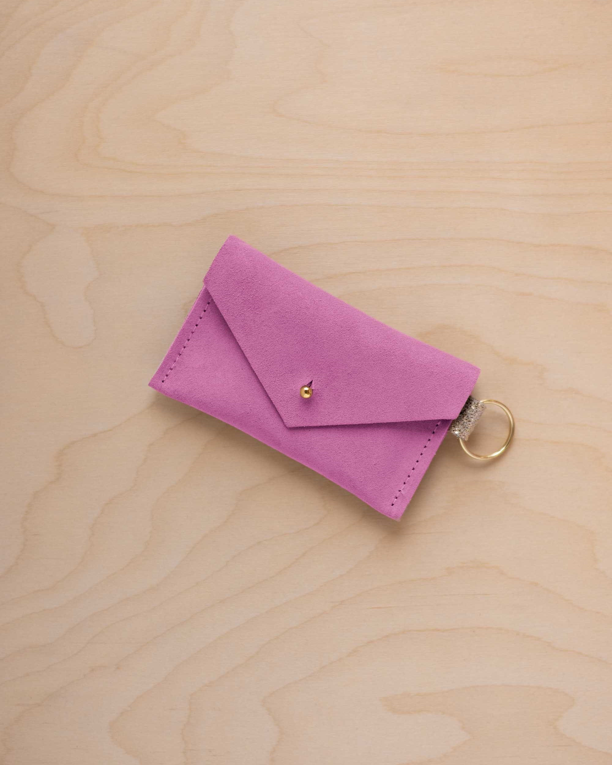 A small peony pink coloured suede envelope-style keyring pouch with a gold button, resting on a light wood grain surface.