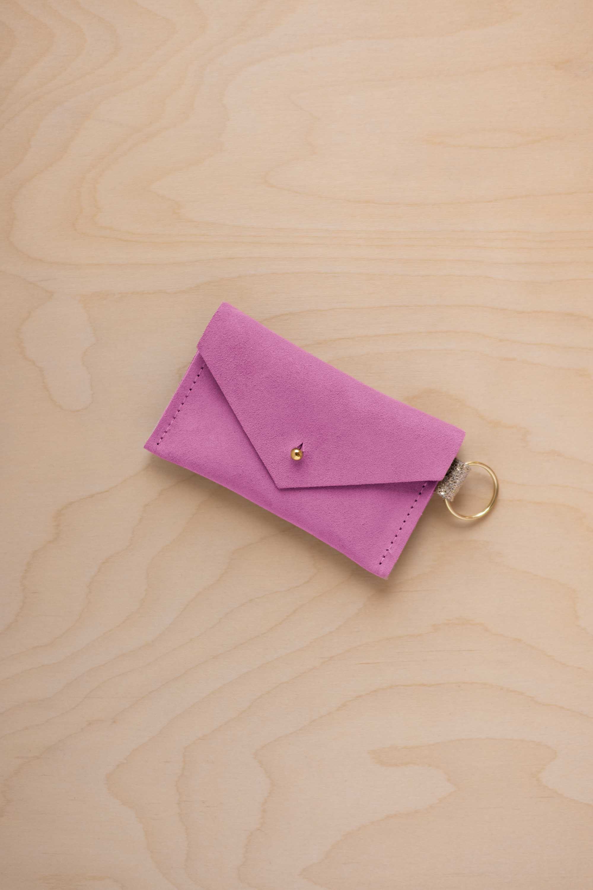 A small peony pink coloured suede envelope-style keyring pouch with a gold button, resting on a light wood grain surface.