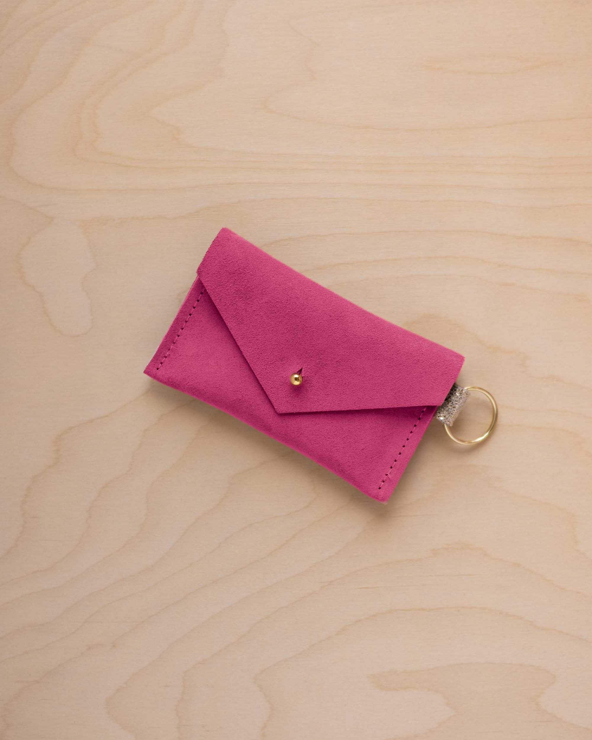 A small raspberry coloured suede envelope-style keyring pouch with a gold button, resting on a light wood grain surface.