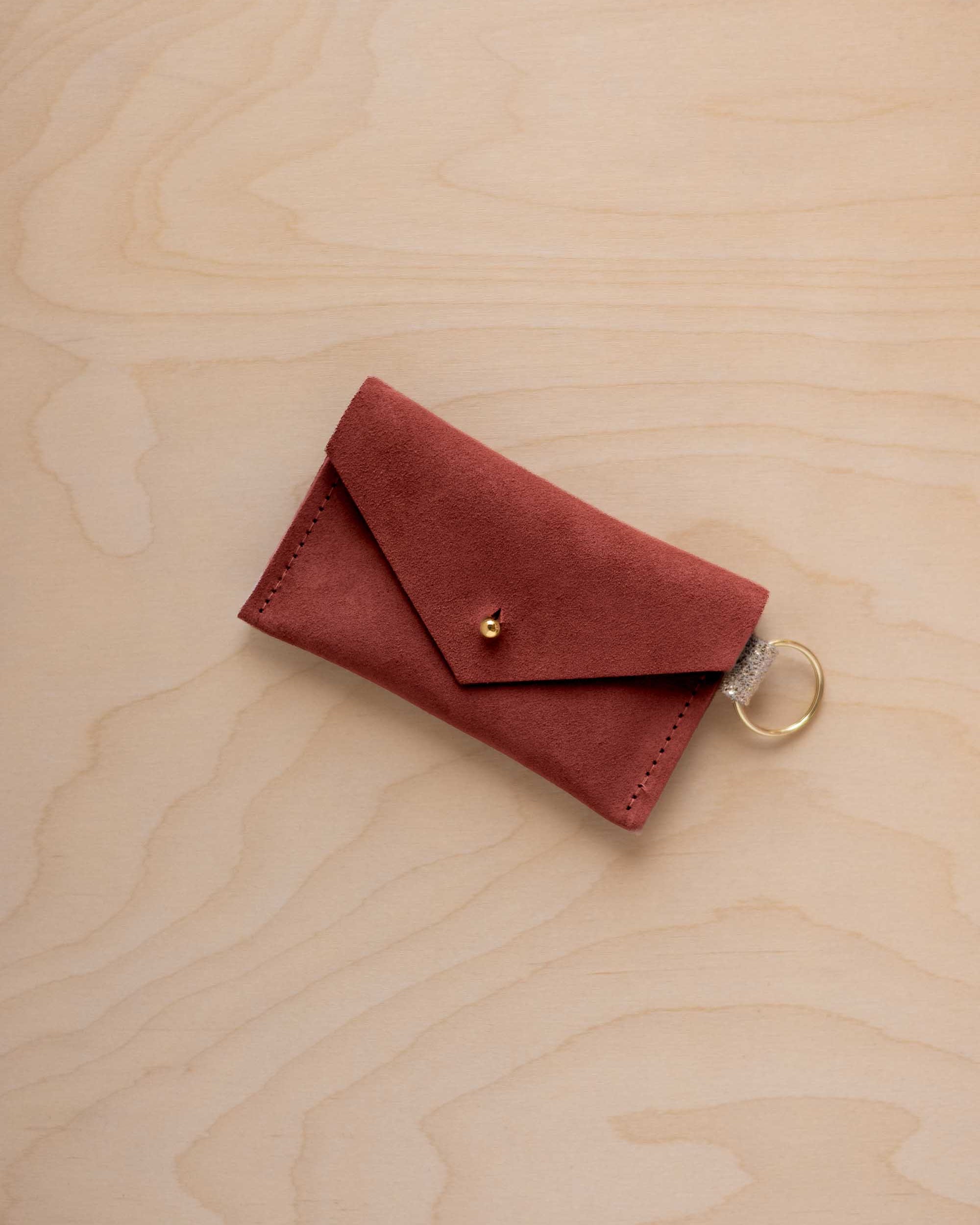 A small rust red coloured suede envelope-style keyring pouch with a gold button, resting on a light wood grain surface.