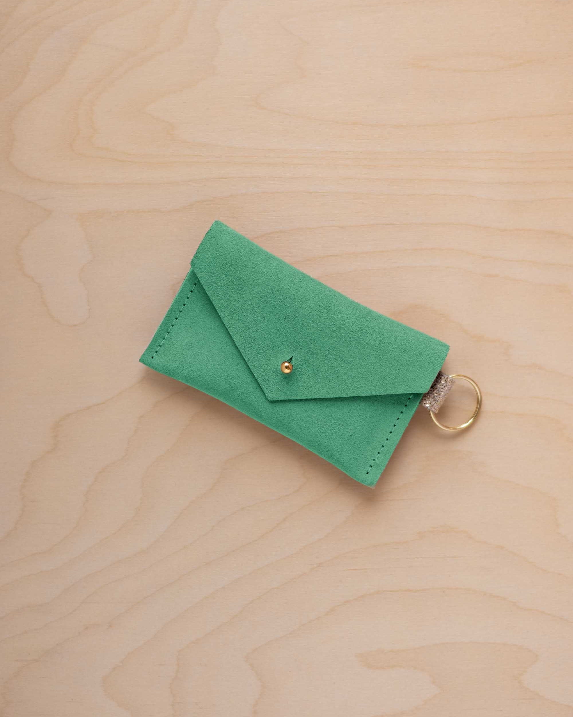 A small seafoam green coloured suede envelope-style keyring pouch with a gold button, resting on a light wood grain surface.