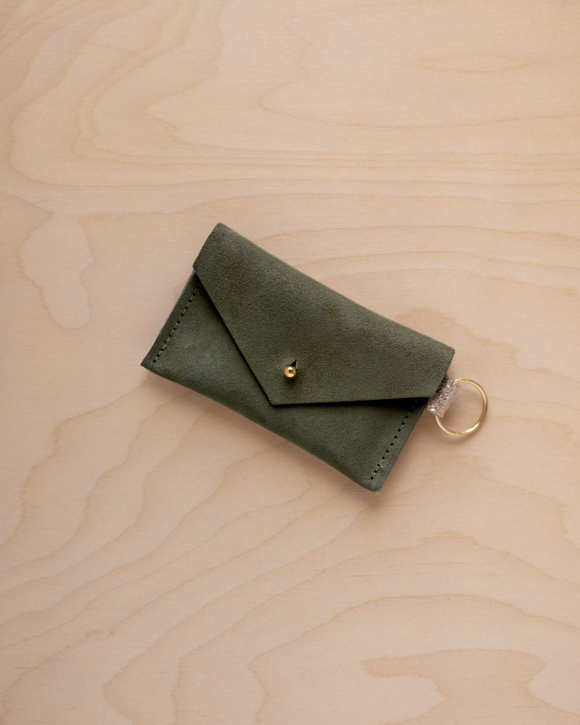 A small soft olive green coloured suede envelope-style keyring pouch with a gold button, resting on a light wood grain surface.