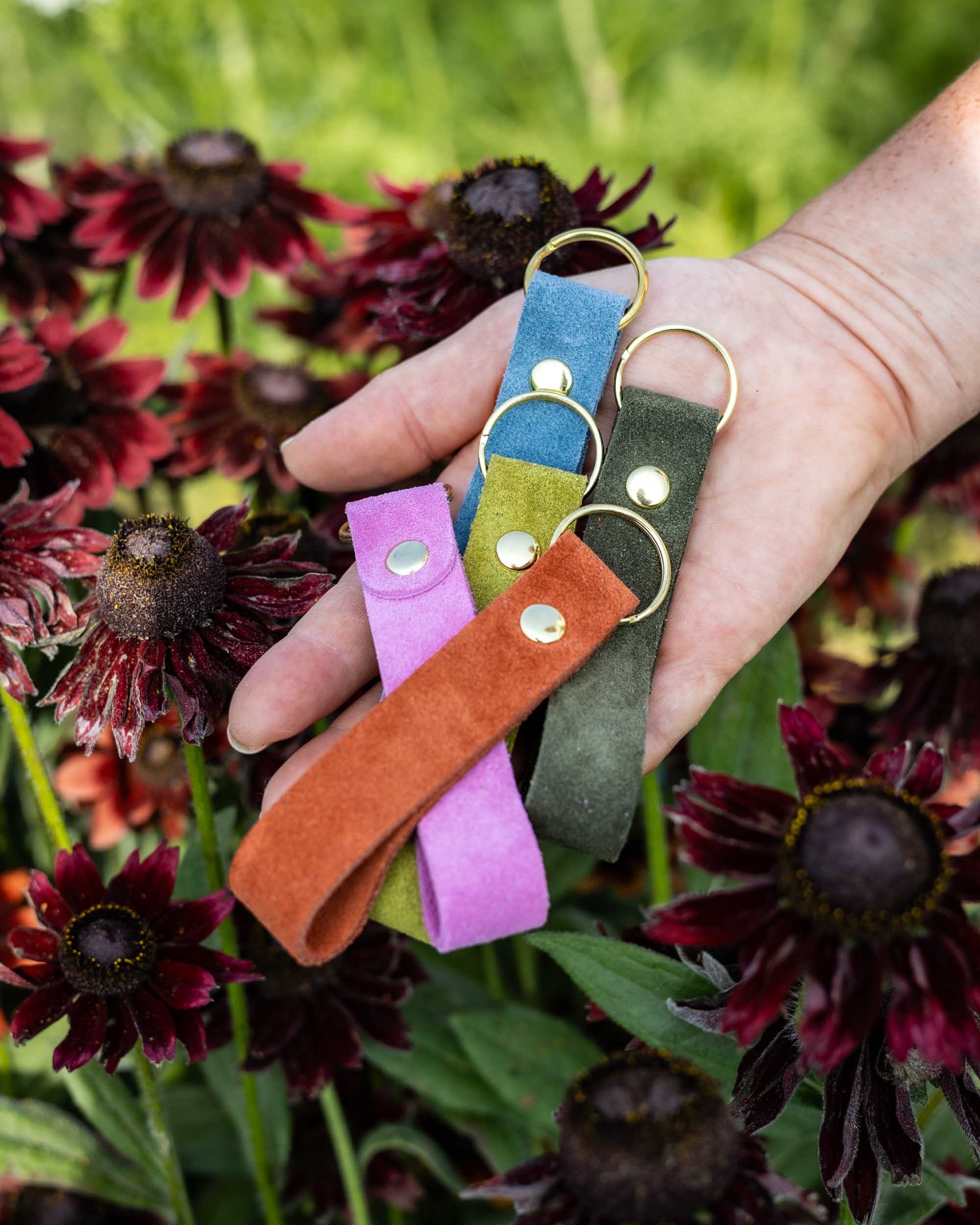 A hand holding five colourful suede keychains (pink, blue, lime green, olive green and terracotta) with gold-coloured metal keyrings, set against a background of dark red coneflowers and green foliage.