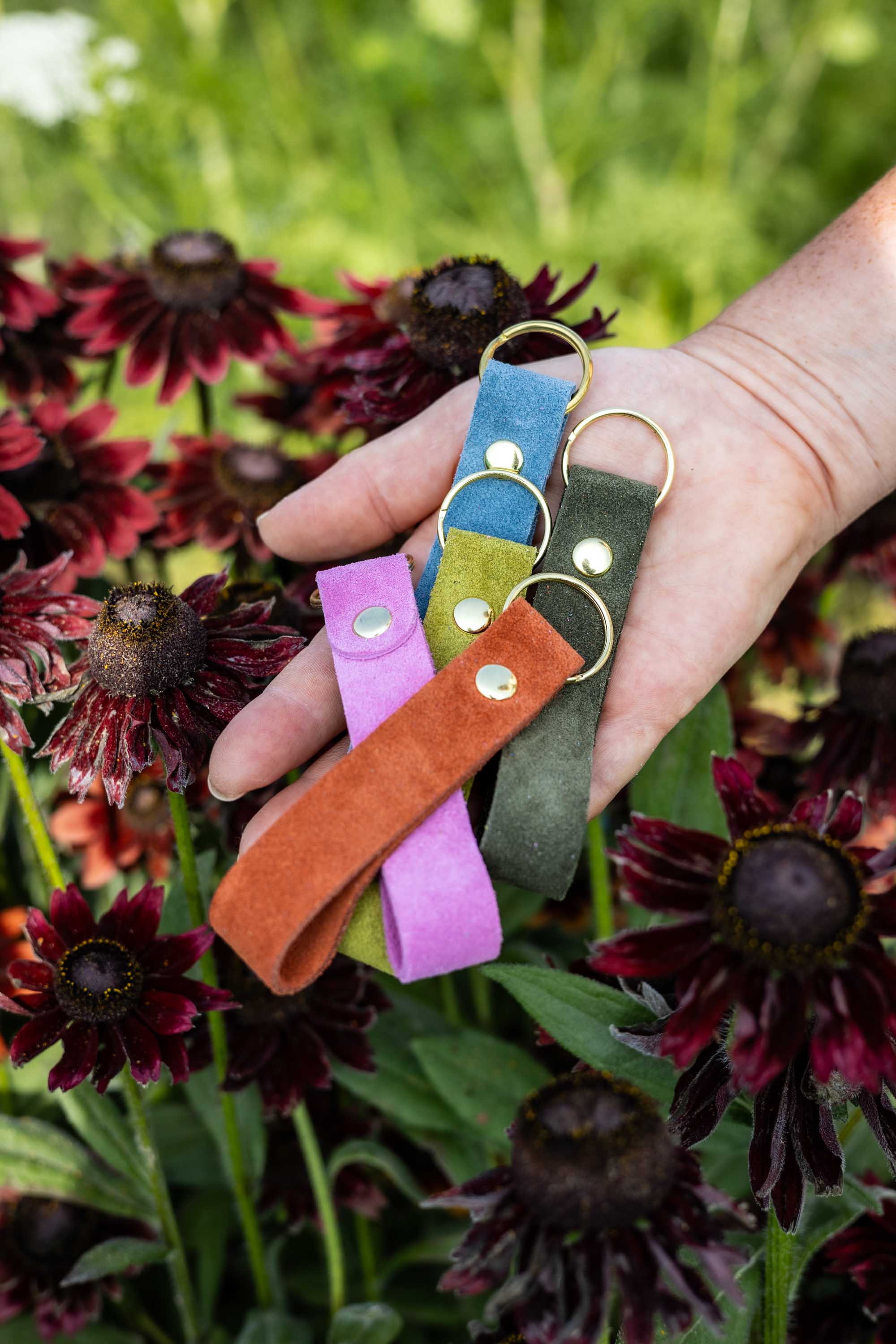 A hand holding five colourful suede keychains (pink, blue, lime green, olive green and terracotta) with gold-coloured metal keyrings, set against a background of dark red coneflowers and green foliage.