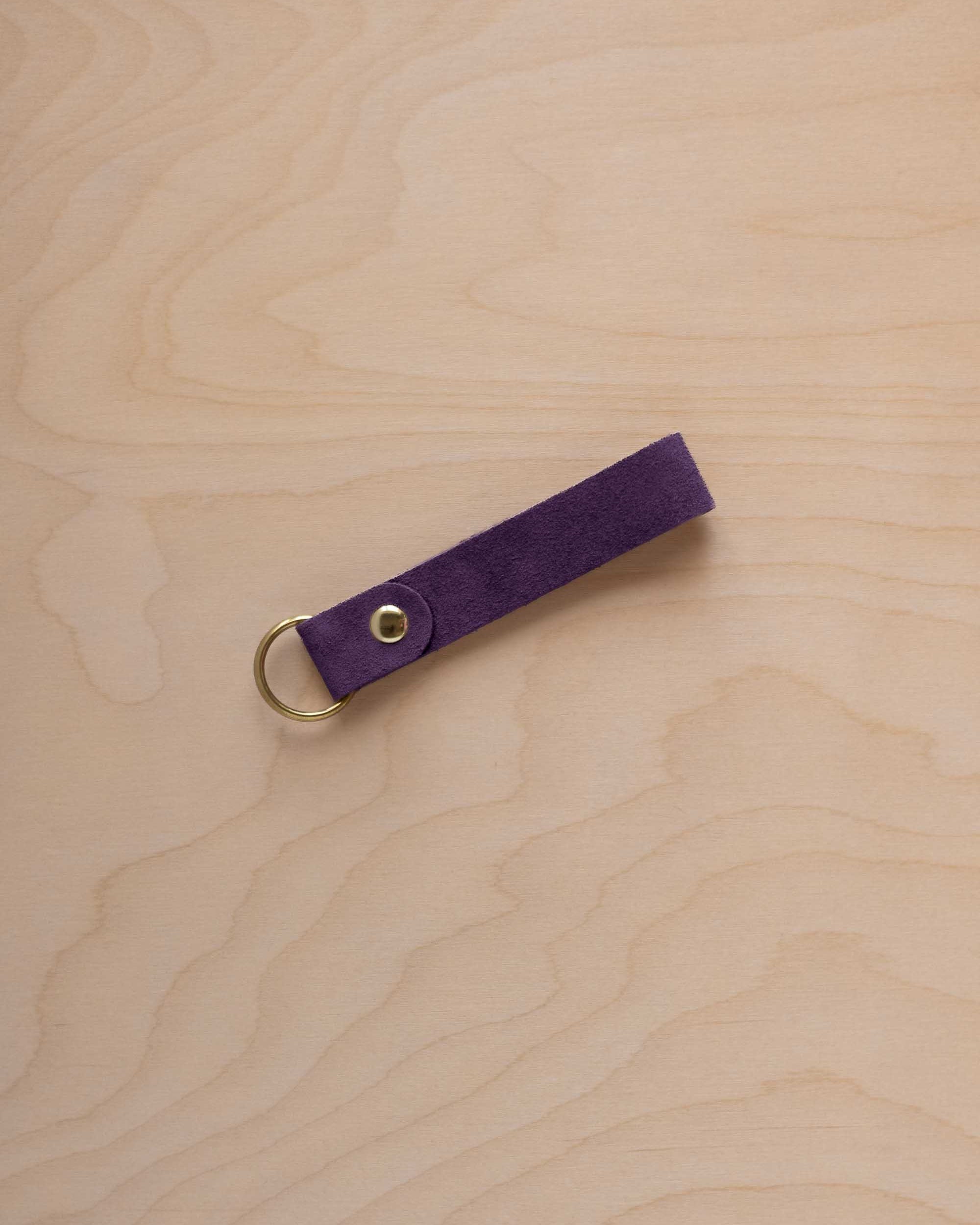 A blackcurrant dark purple coloured suede leather keychain with a gold metal ring and rivet, displayed diagonally on a light wood grain background.