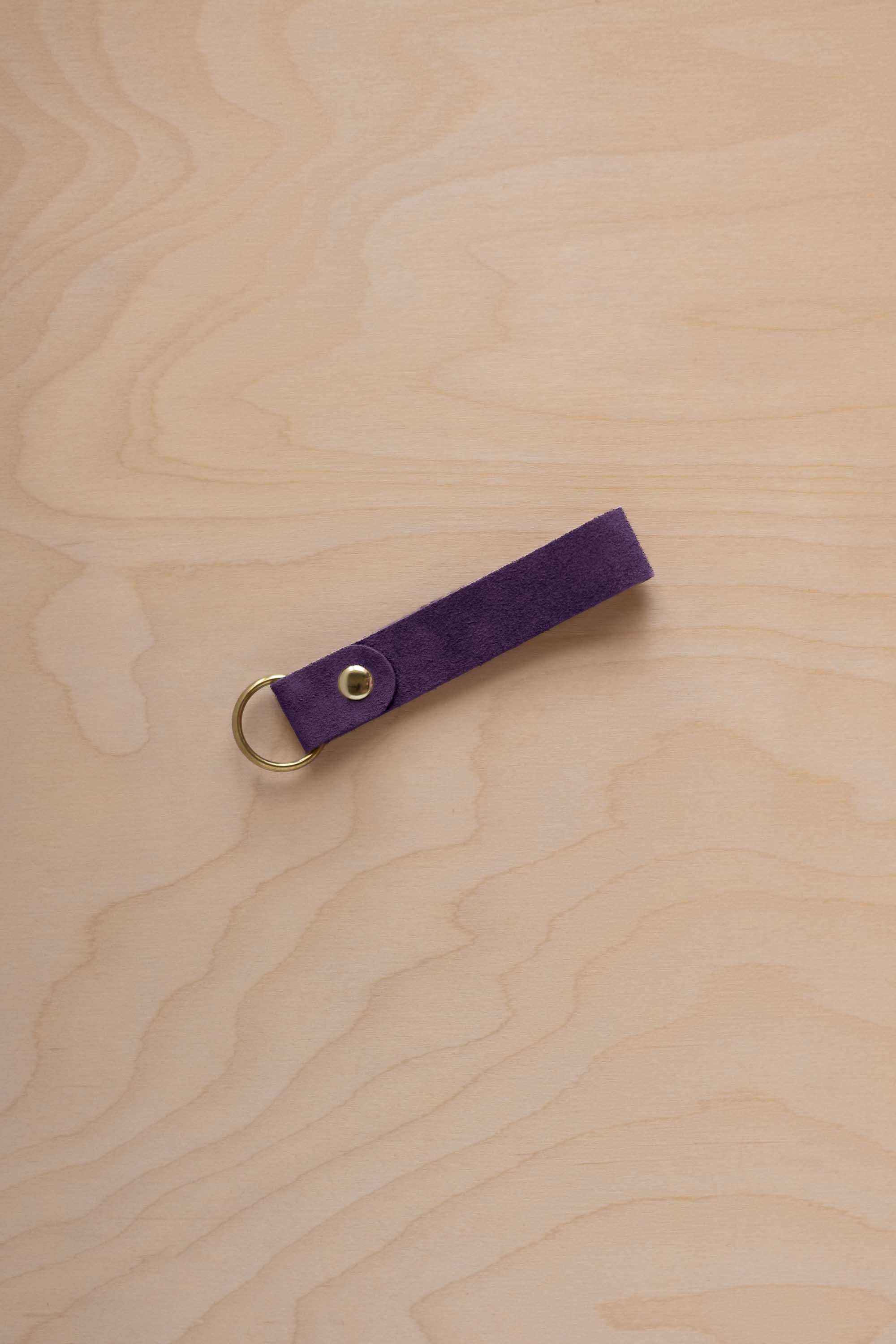 A blackcurrant dark purple coloured suede leather keychain with a gold metal ring and rivet, displayed diagonally on a light wood grain background.