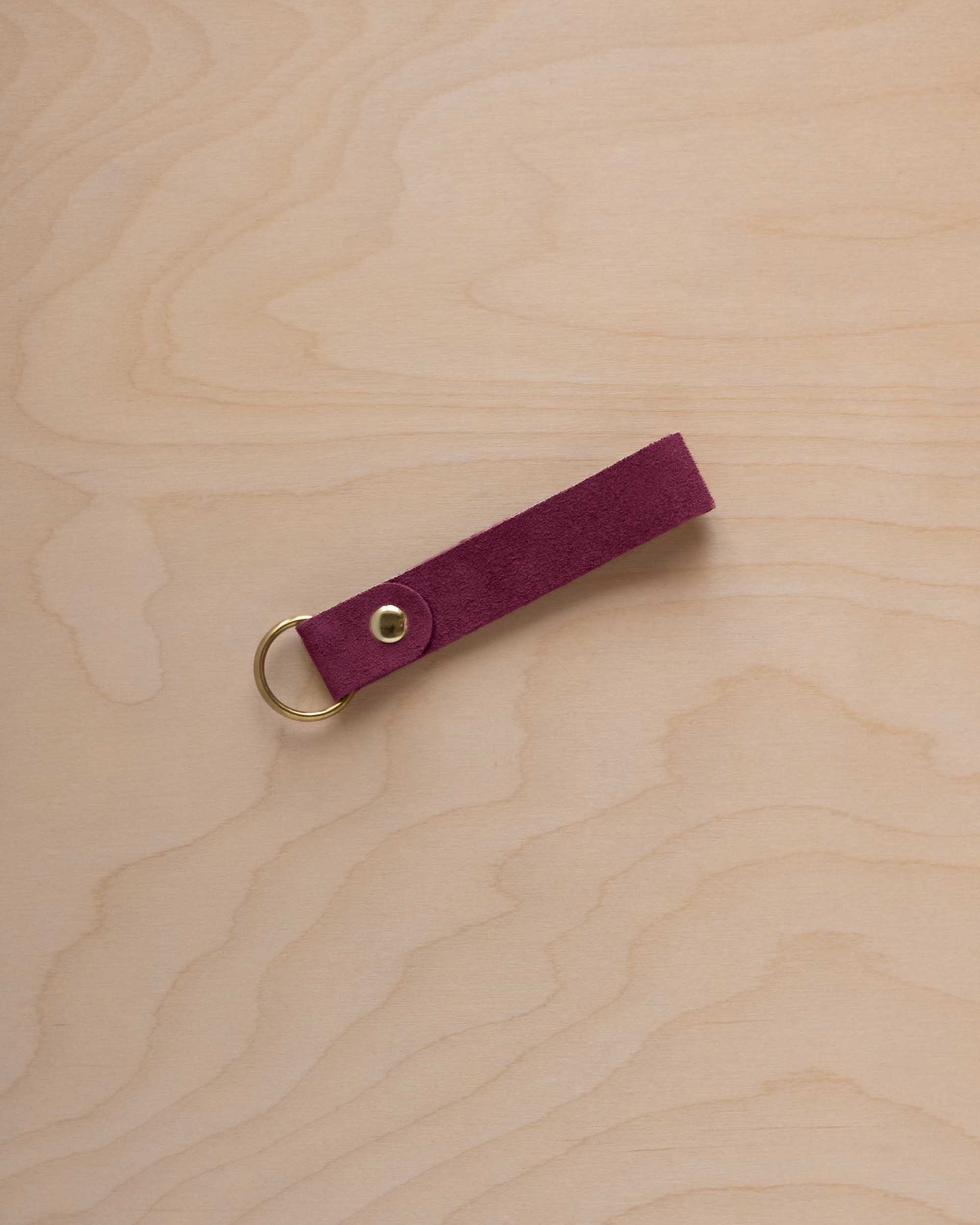 A burgundy suede leather keychain with a gold metal ring and rivet, displayed diagonally on a light wood grain background.