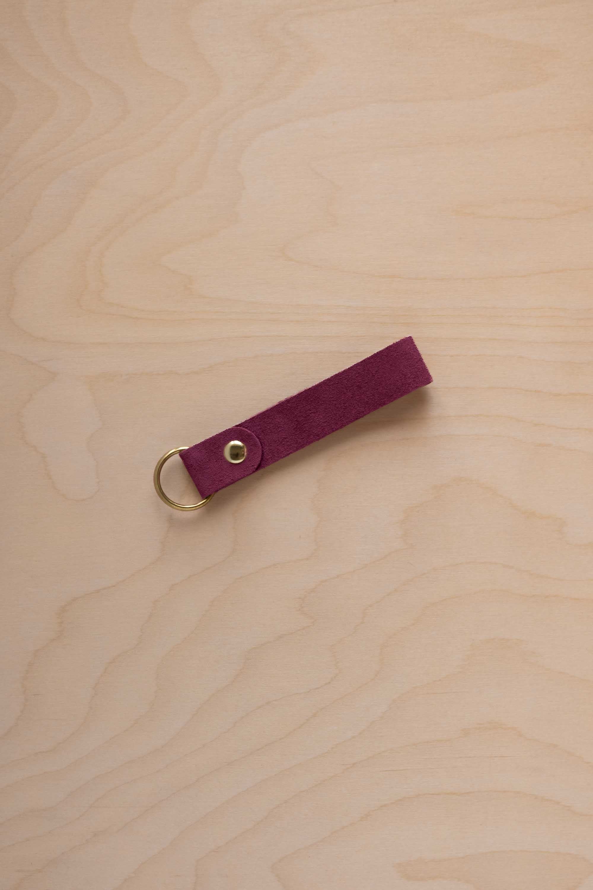 A burgundy suede leather keychain with a gold metal ring and rivet, displayed diagonally on a light wood grain background.