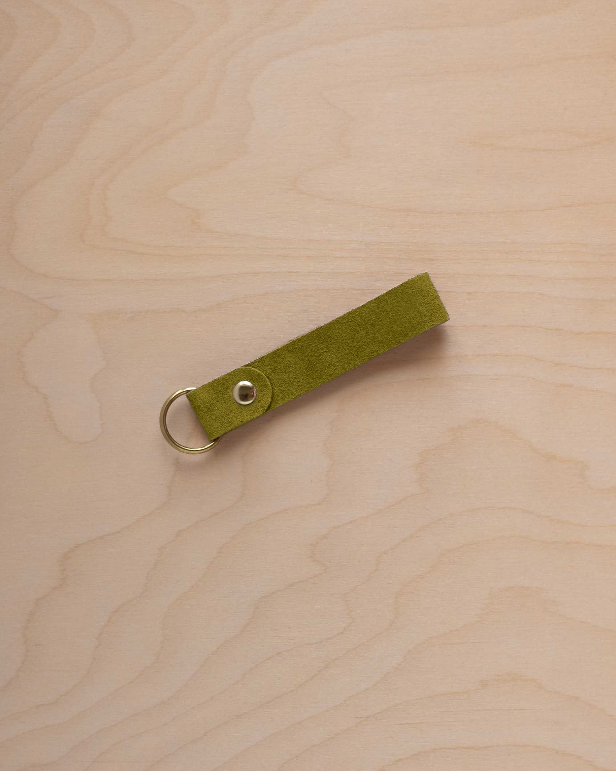 A chartreuse lime green coloured suede leather keychain with a gold metal ring and rivet, displayed diagonally on a light wood grain background.