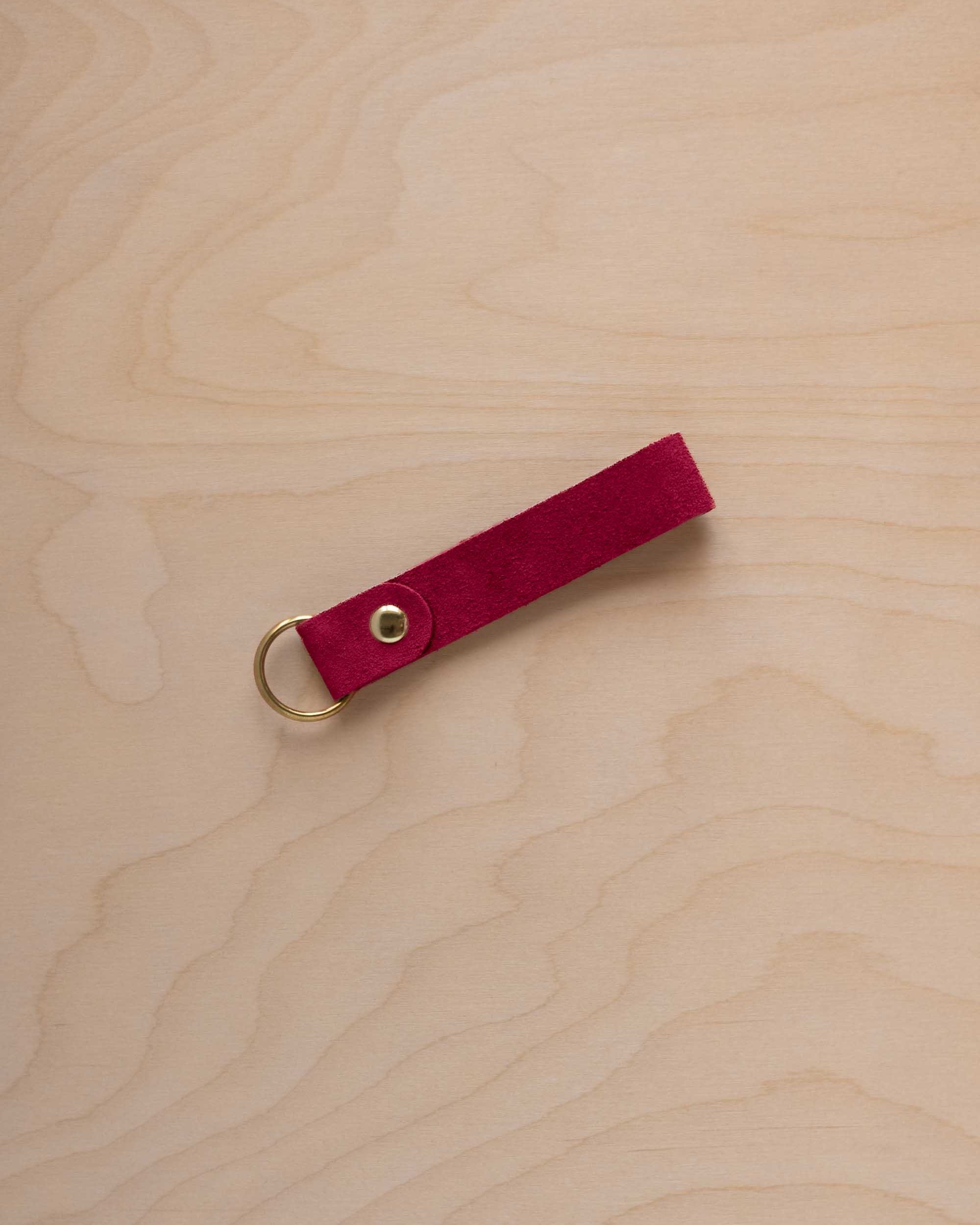 A crimson red suede leather keychain with a gold metal ring and rivet, displayed diagonally on a light wood grain background.