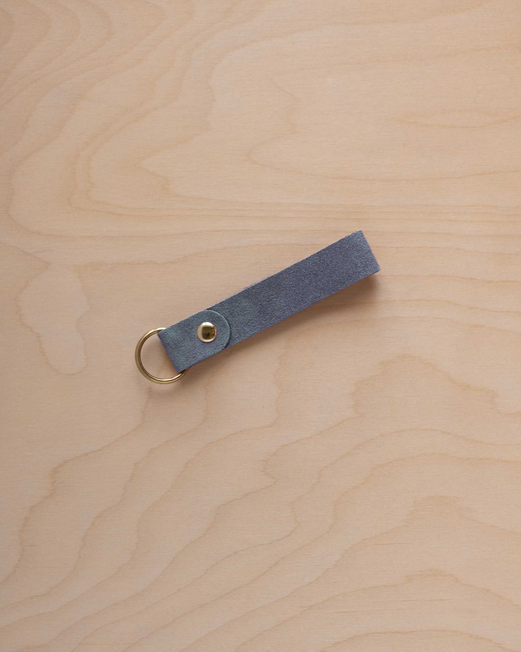 A dusky blue suede leather keychain with a gold metal ring and rivet, displayed diagonally on a light wood grain background.