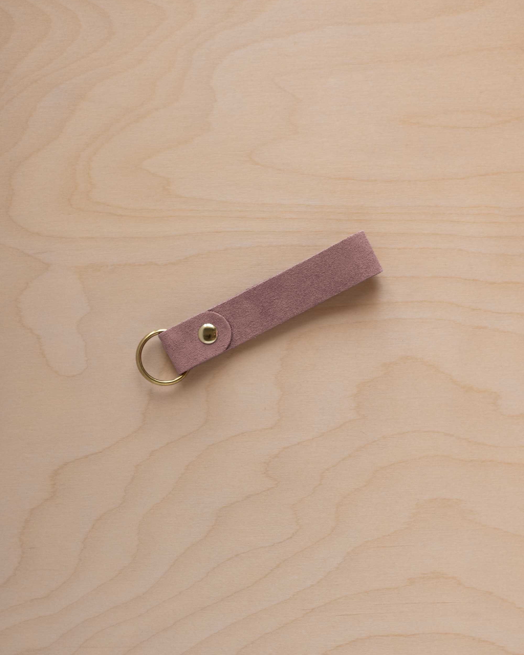 A dusty mauve suede leather keychain with a gold metal ring and rivet, displayed diagonally on a light wood grain background.