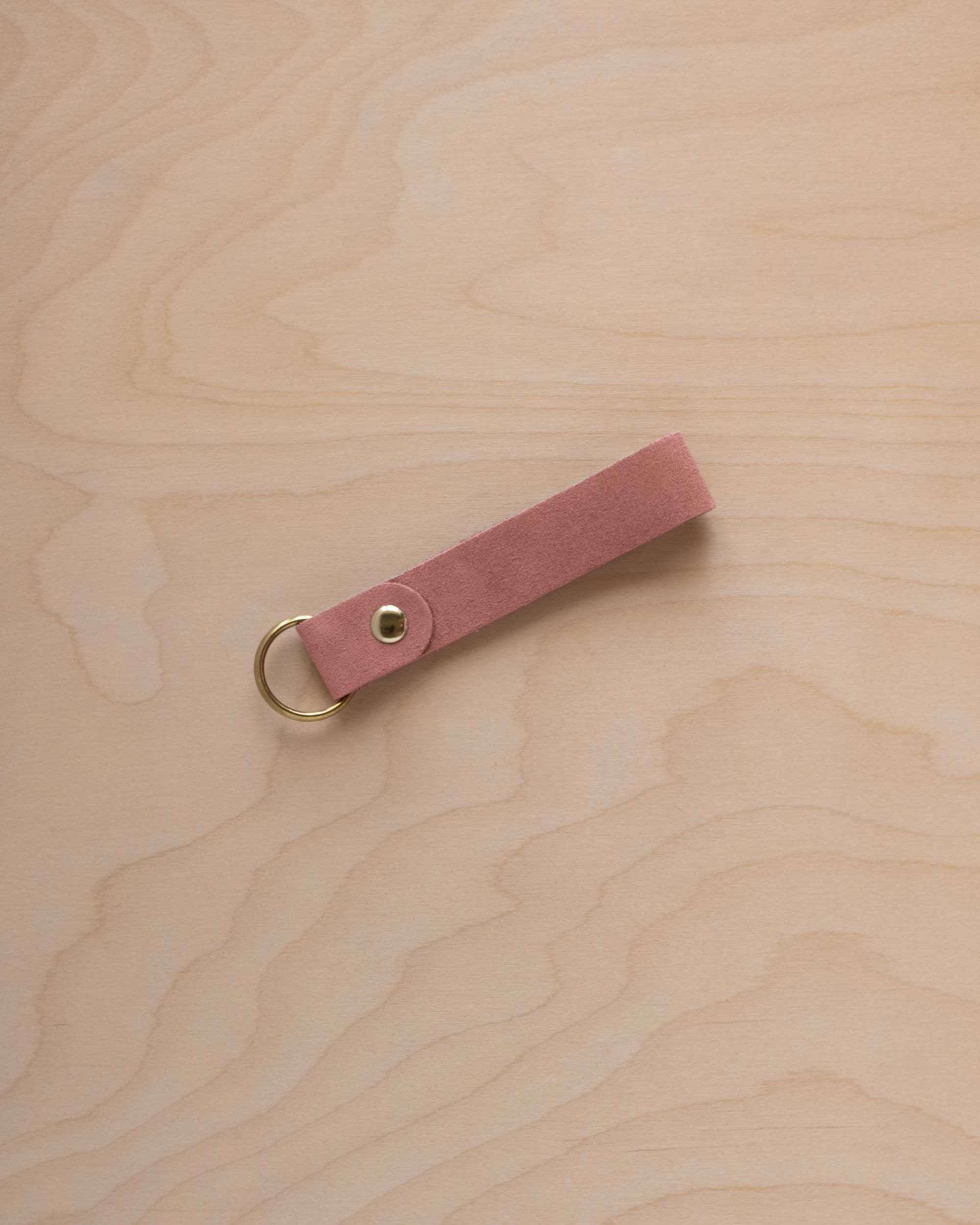 A dusty rose pale pink coloured suede leather keychain with a gold metal ring and rivet, displayed diagonally on a light wood grain background.