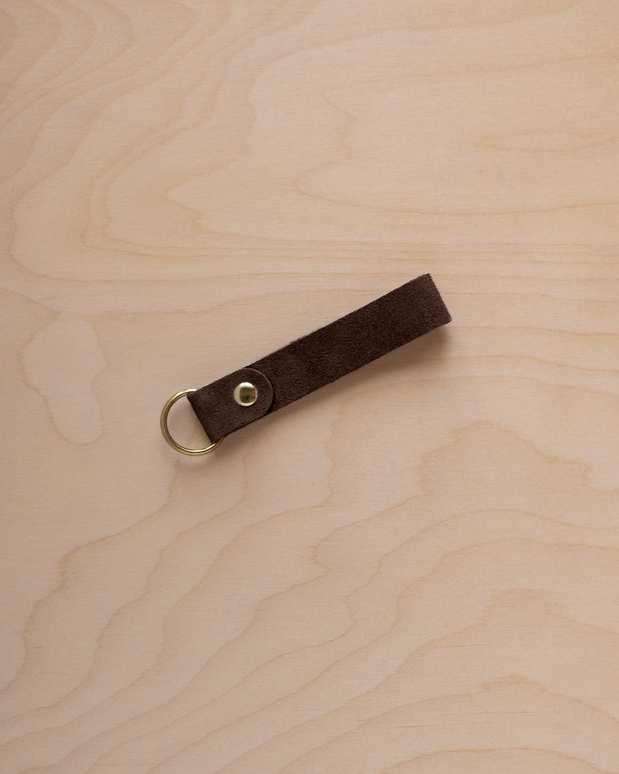 An espresso brown suede leather keychain with a gold metal ring and rivet, displayed diagonally on a light wood grain background.