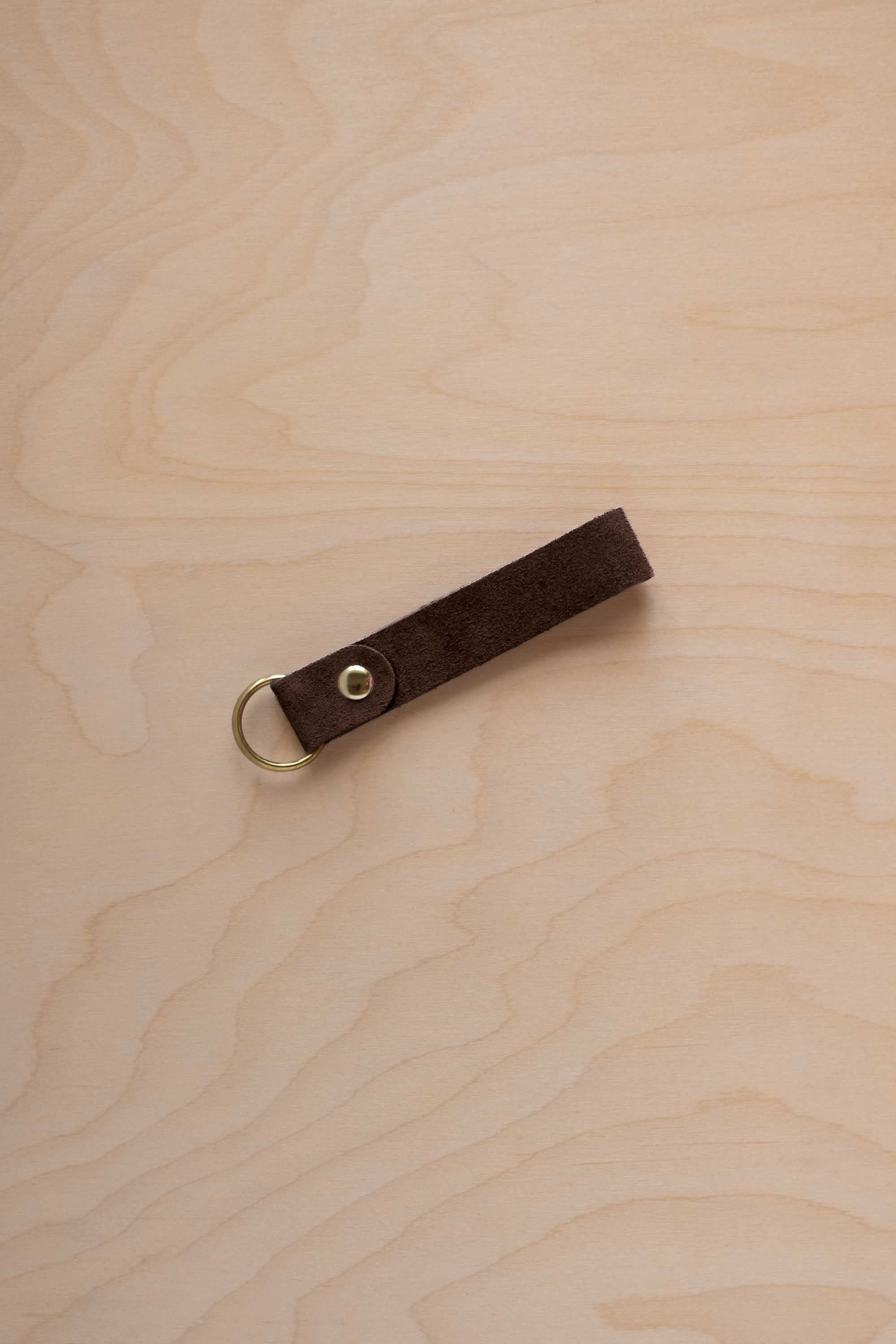An espresso brown suede leather keychain with a gold metal ring and rivet, displayed diagonally on a light wood grain background.