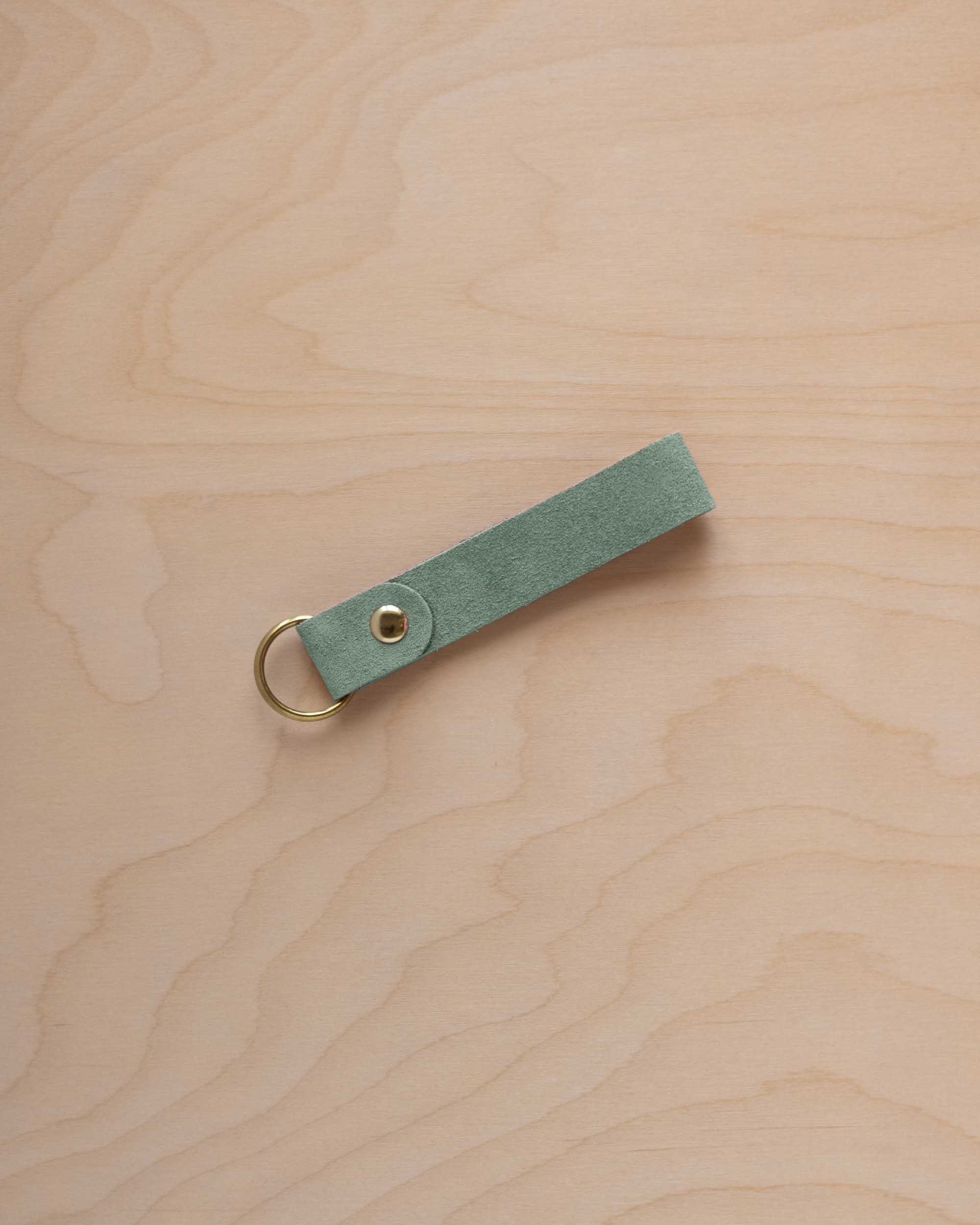 A eucalyptus mint green coloured suede leather keychain with a gold metal ring and rivet, displayed diagonally on a light wood grain background.