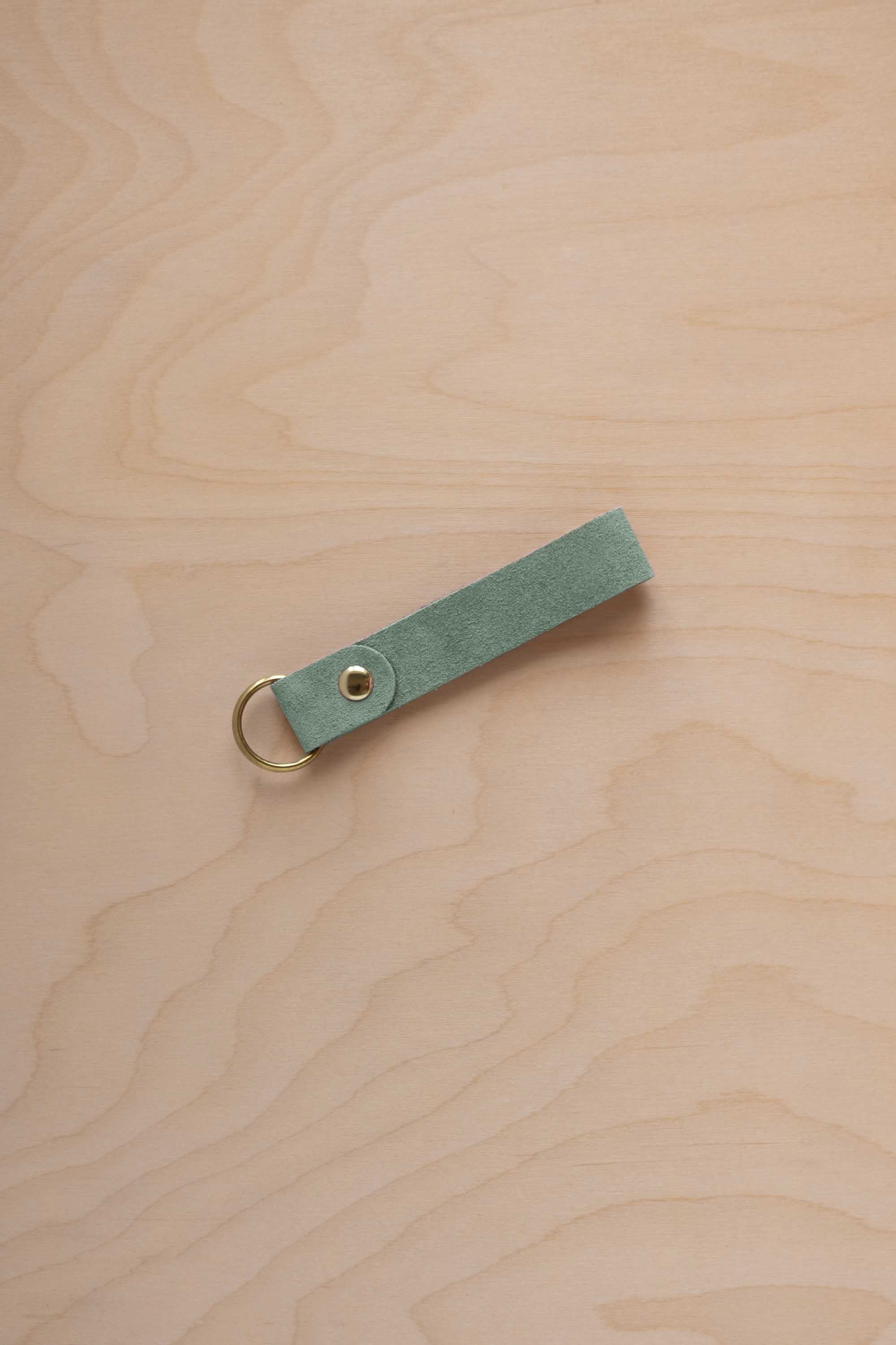 A eucalyptus mint green coloured suede leather keychain with a gold metal ring and rivet, displayed diagonally on a light wood grain background.