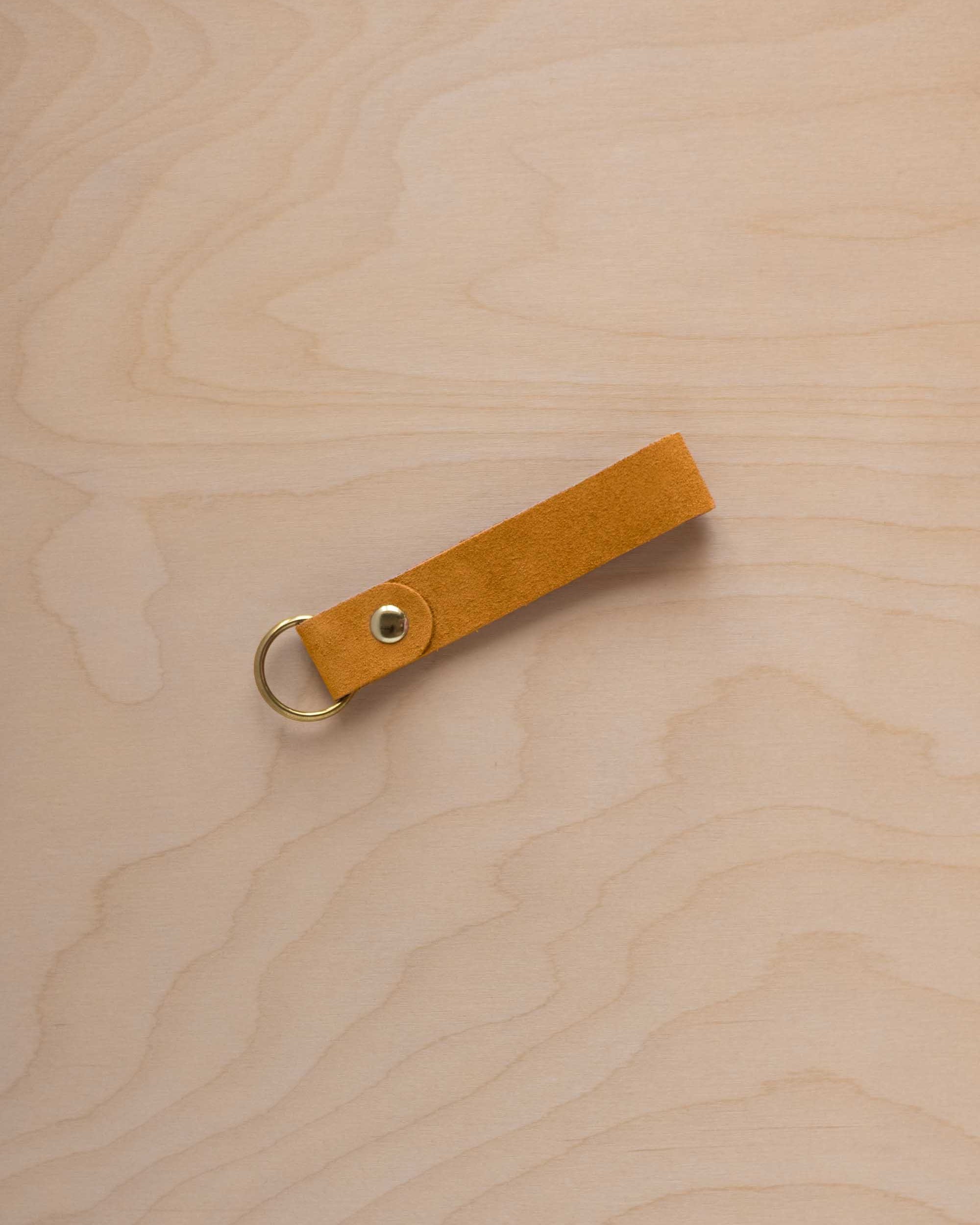 A golden ochre mustard yellow coloured beige suede leather keychain with a gold metal ring and rivet, displayed diagonally on a light wood grain background.