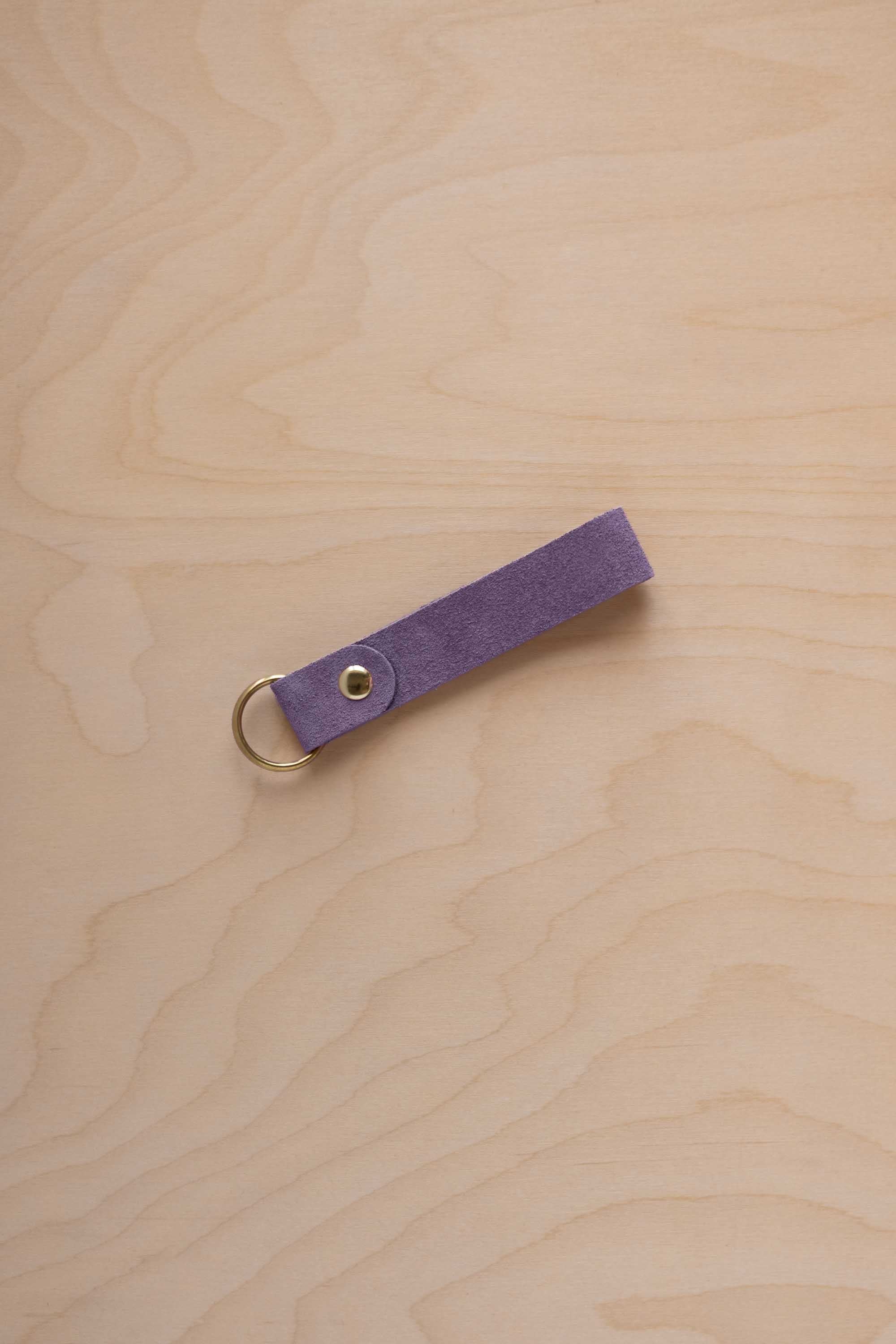 A lavender coloured suede leather keychain with a gold metal ring and rivet, displayed diagonally on a light wood grain background.