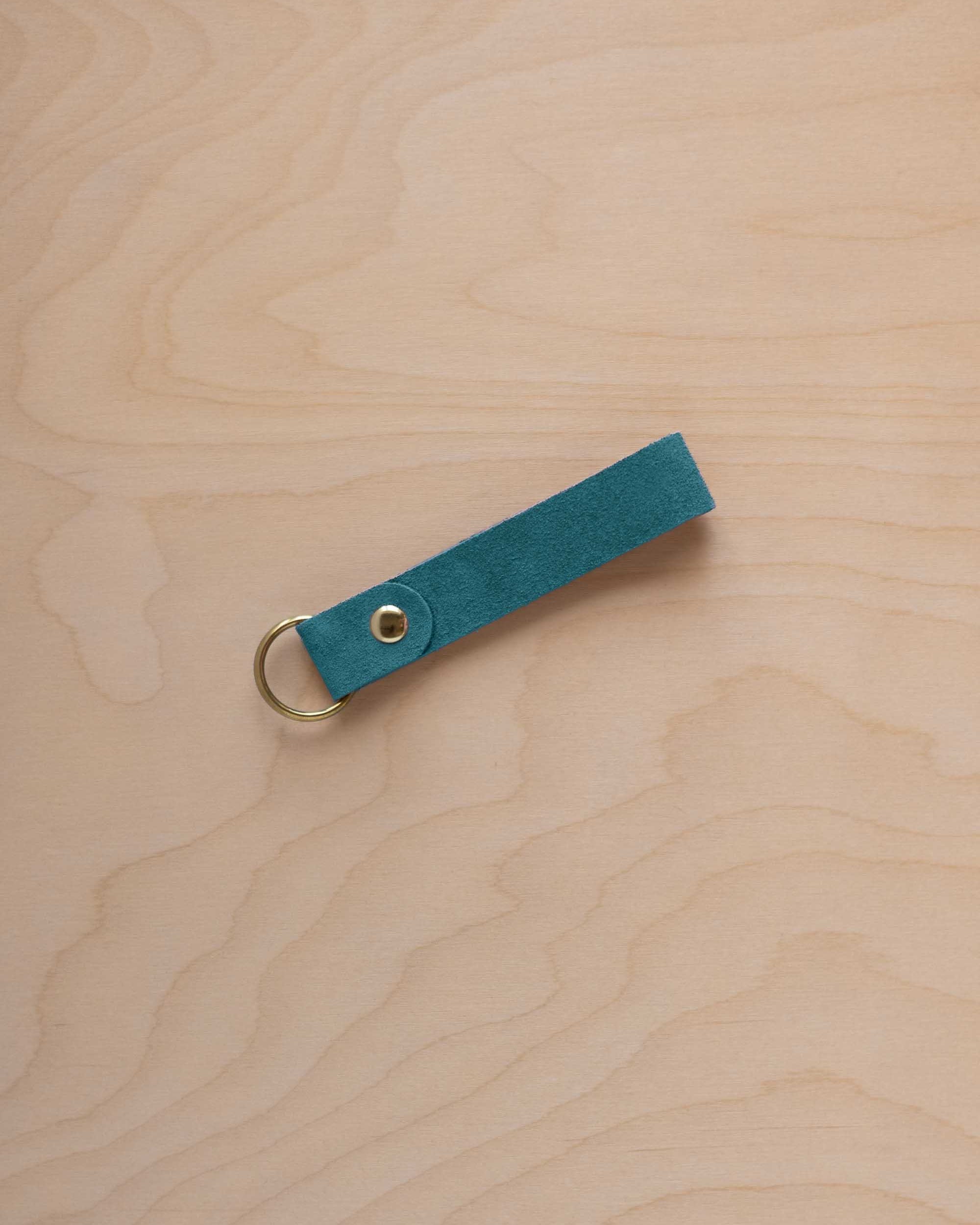 A marine teal coloured suede leather keychain with a gold metal ring and rivet, displayed diagonally on a light wood grain background.