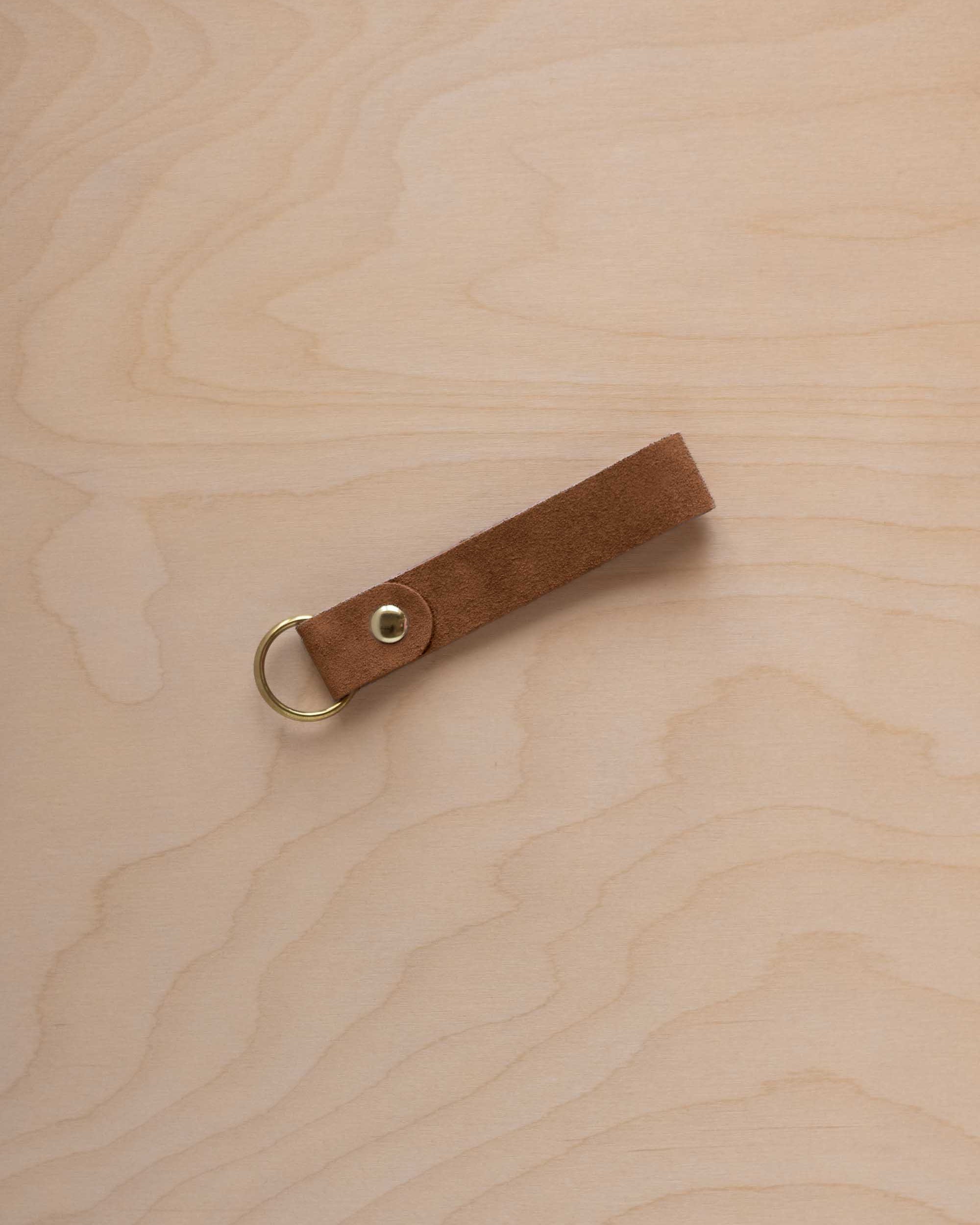 A mocha brown suede leather keychain with a gold metal ring and rivet, displayed diagonally on a light wood grain background.