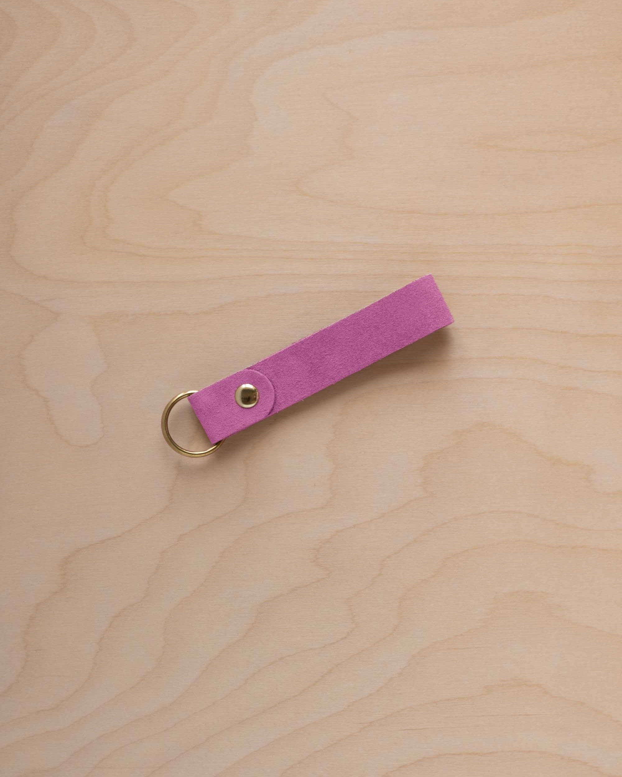A peony pink suede leather keychain with a gold metal ring and rivet, displayed diagonally on a light wood grain background.