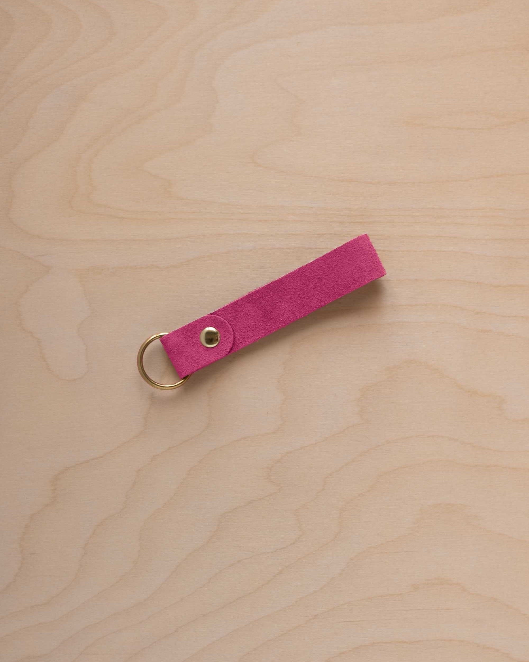 A raspberry pink suede leather keychain with a gold metal ring and rivet, displayed diagonally on a light wood grain background.