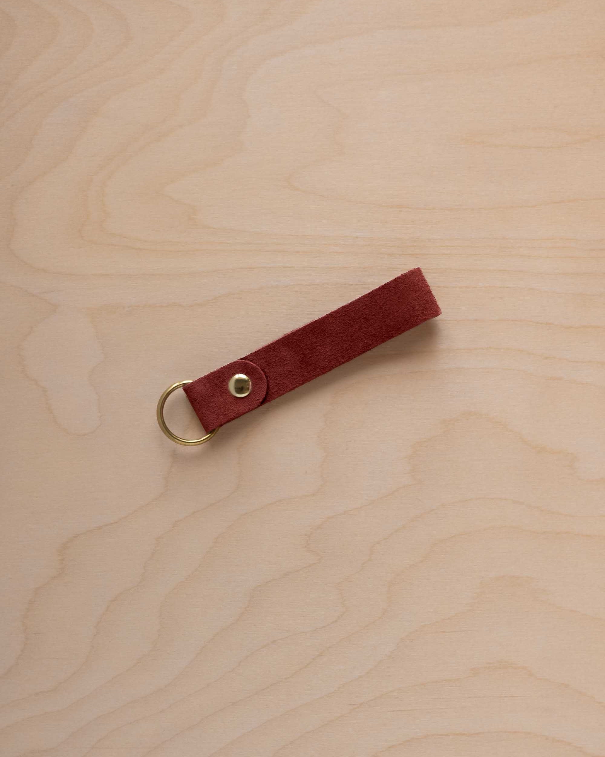 A rust red suede leather keychain with a gold metal ring and rivet, displayed diagonally on a light wood grain background.