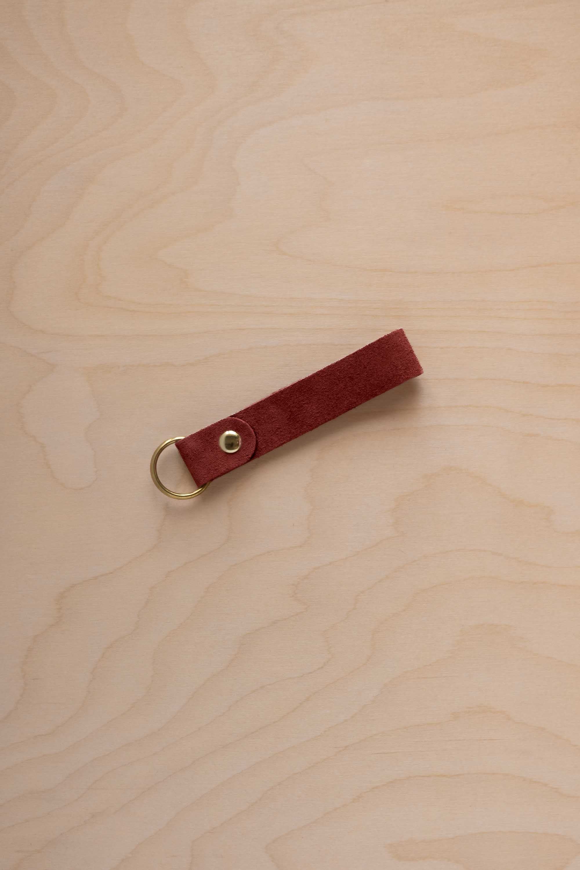 A rust red suede leather keychain with a gold metal ring and rivet, displayed diagonally on a light wood grain background.