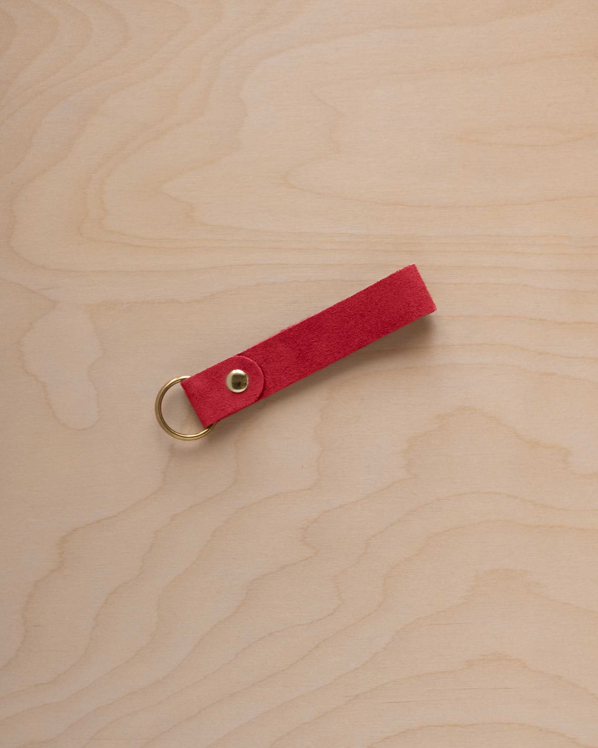 A scarlet red suede leather keychain with a gold metal ring and rivet, displayed diagonally on a light wood grain background.
