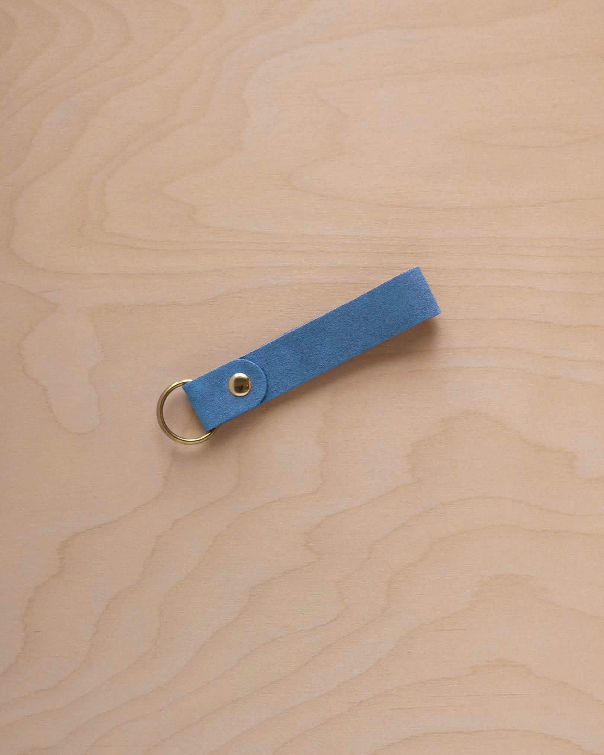 A sky blue suede leather keychain with a gold metal ring and rivet, displayed diagonally on a light wood grain background.